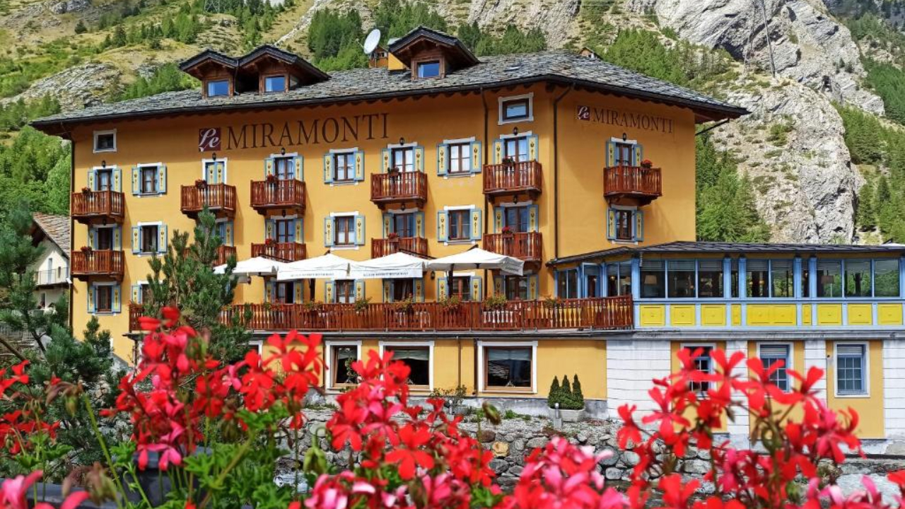 Immerso nell'incantevole villaggio di La Thuile, questo pittoresco hotel giallo presenta balconi in legno ed è circondato da una vegetazione lussureggiante e da vivaci fiori rossi. Adagiato su uno sfondo di montagne aspre, tutto si svolge sotto un limpido cielo azzurro.