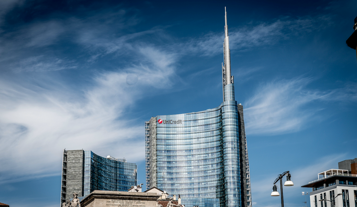 Un moderno grattacielo con un design curvo unico e una guglia appuntita si staglia contro un cielo azzurro con nuvole vaporose. L'edificio di UniCredit ha finestre in vetro riflettente ed è circondato da alcune strutture più piccole e da un lampione in primo piano.