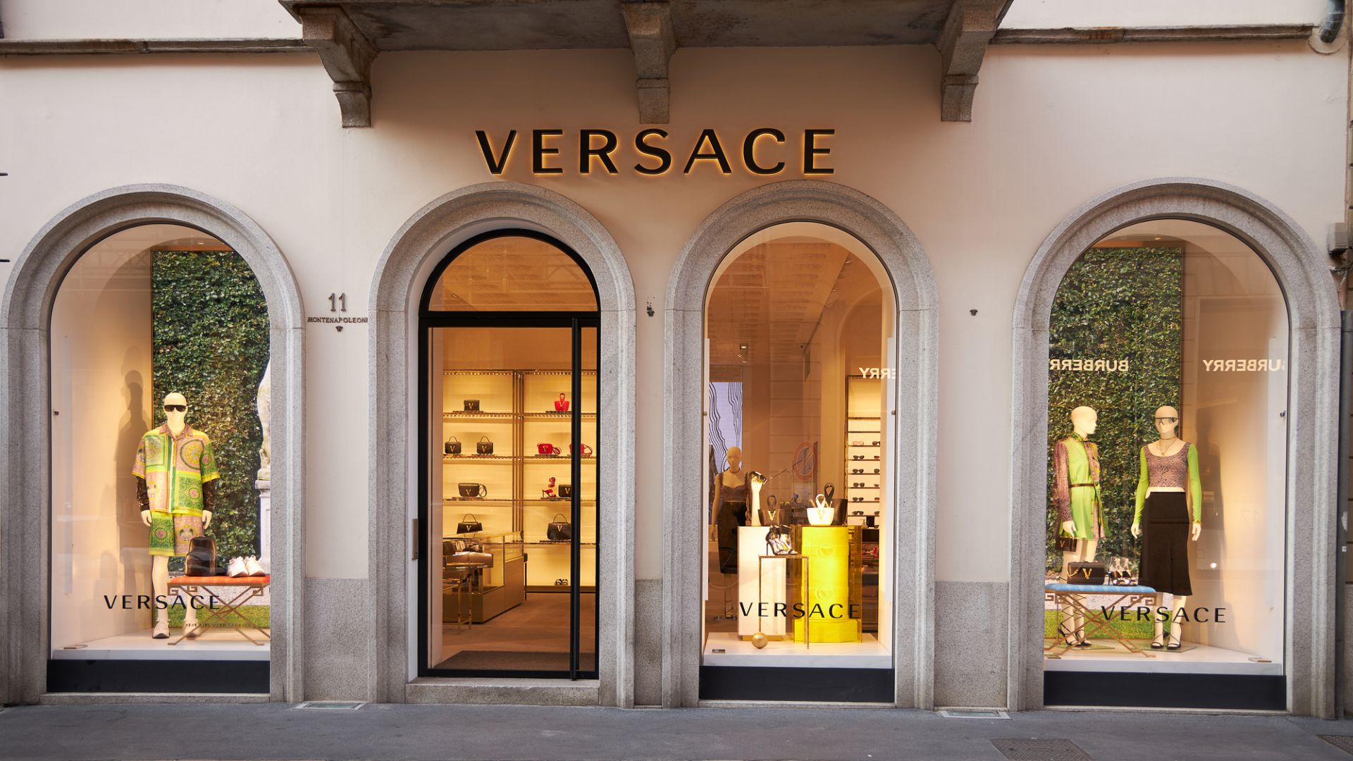 Un negozio Versace con tre grandi vetrine ad arco che espongono manichini in abiti eleganti. Il nome del negozio è ben visibile in alto. All'interno, sono visibili scaffali con prodotti ordinatamente disposti. La facciata ha un design classico.