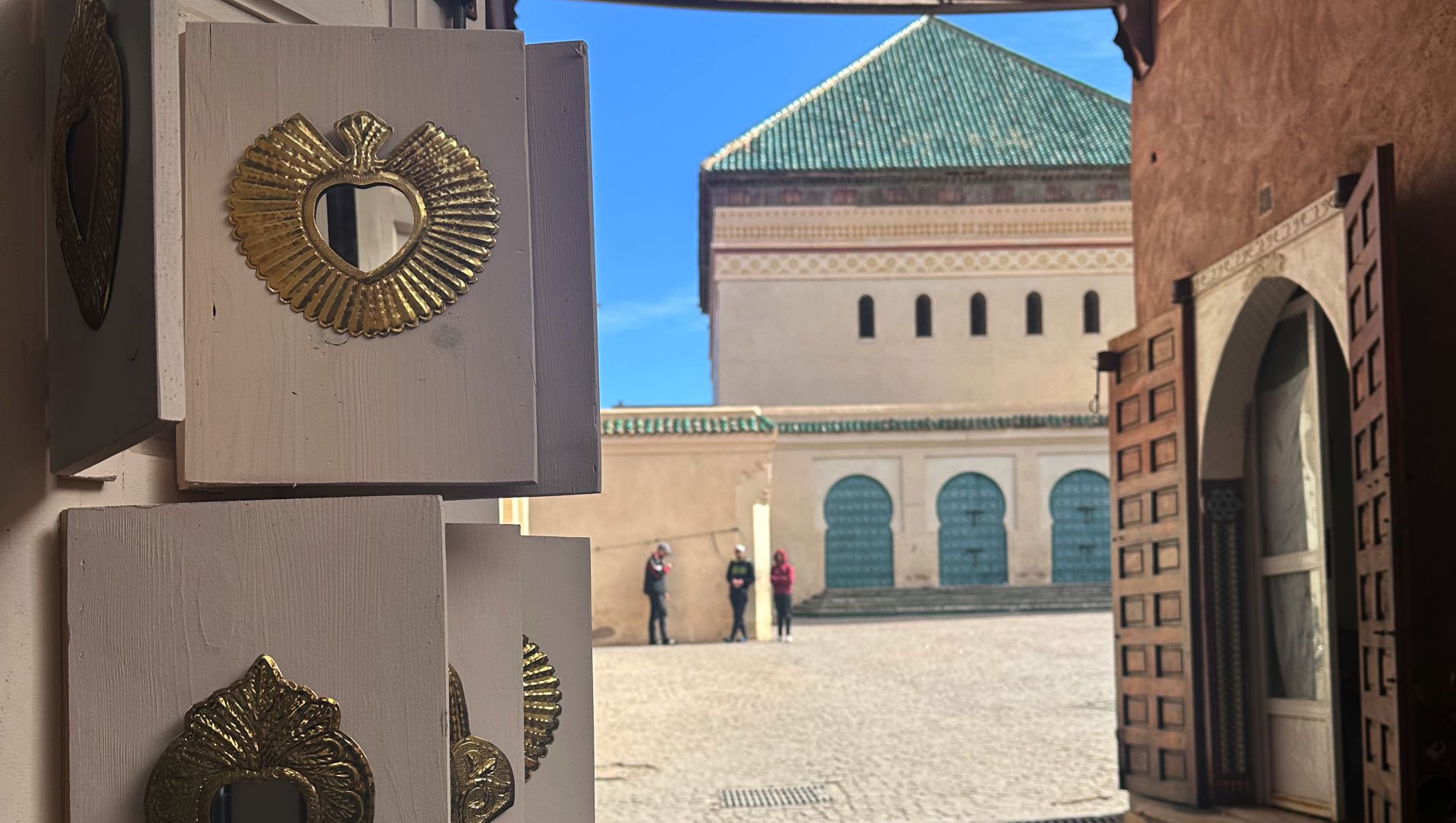 Una vista del cortile incorniciata da un ingresso aperto, con una porta decorata con decorazioni dorate sulla sinistra. Sullo sfondo, un grande edificio con un tetto di tegole verdi e finestre ad arco. Due persone sono in piedi in lontananza.