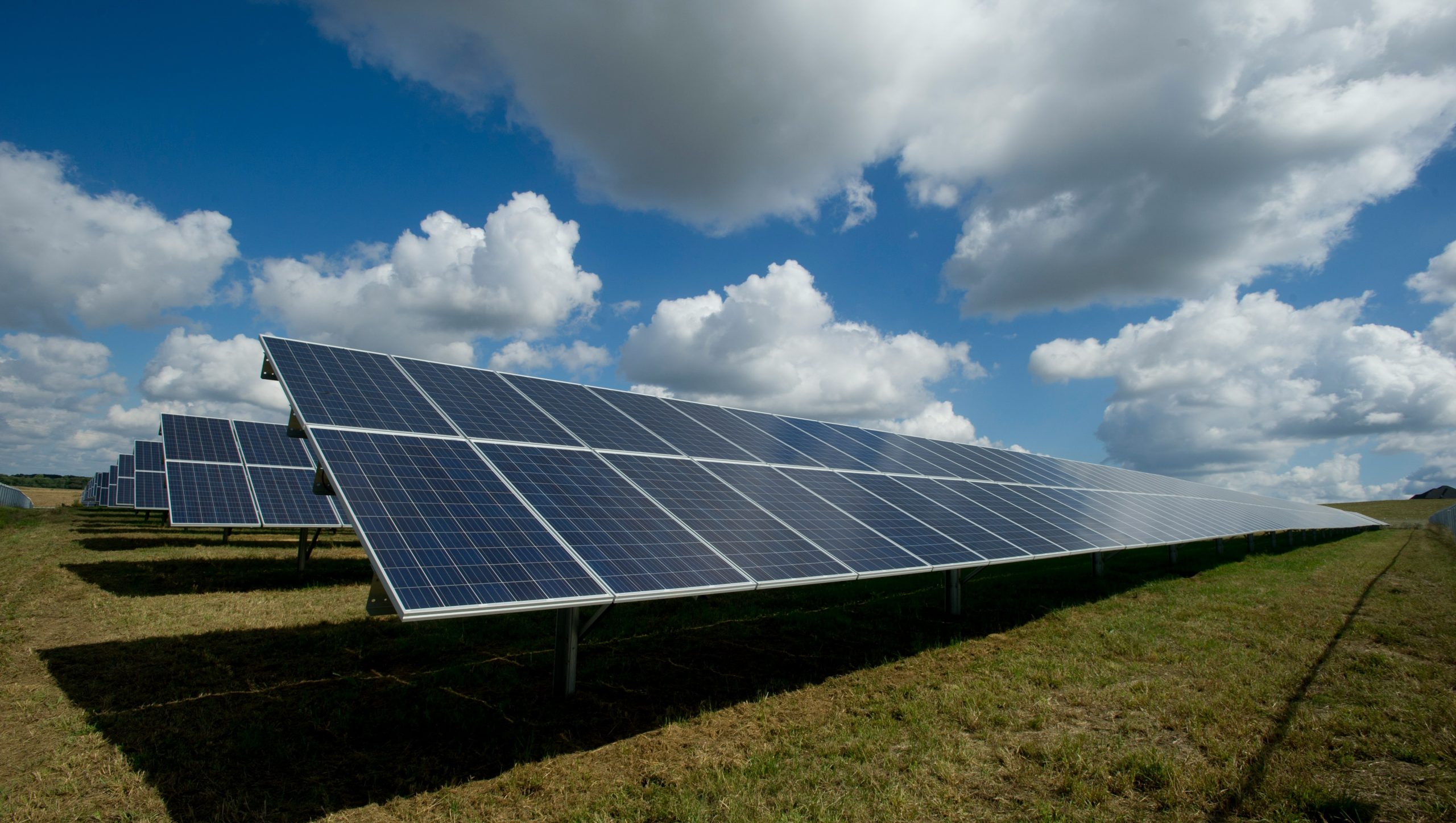 Un campo di pannelli solari sotto un cielo parzialmente nuvoloso simboleggia la transizione energetica. I pannelli sono ordinatamente disposti su un terreno erboso, riflettendo la luce del sole. Soffici nuvole bianche e un cielo azzurro esaltano l'atmosfera luminosa e serena del progresso sostenibile.