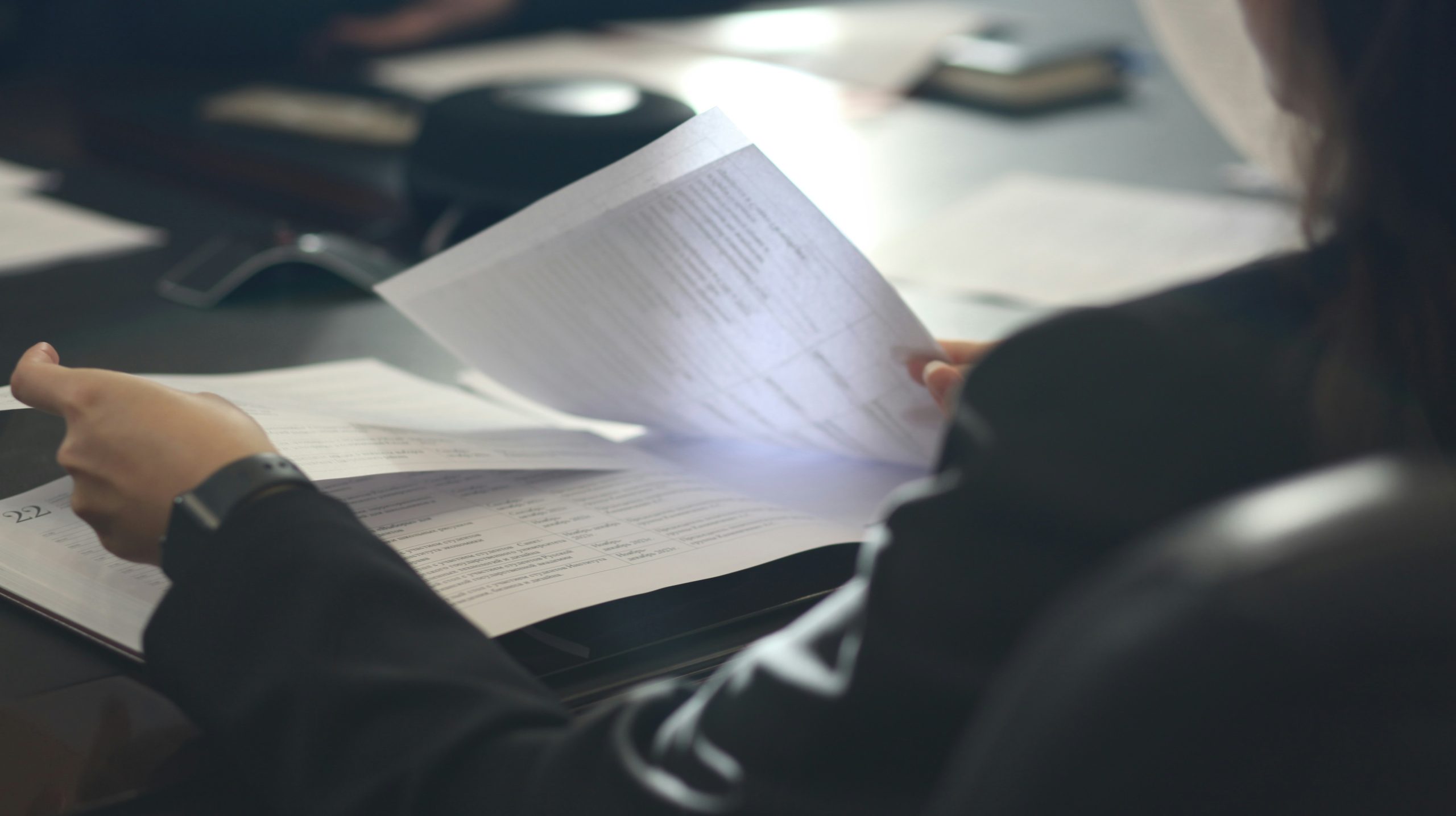 Una persona in tailleur è seduta a un tavolo e sta esaminando documenti stampati, con altri fogli sparsi sulla superficie. L'attenzione è rivolta alle loro mani e ai loro documenti, mentre si addentrano nelle polizze assicurative sulla vita all'estero per le aziende, trasmettendo un senso di lavoro o studio.