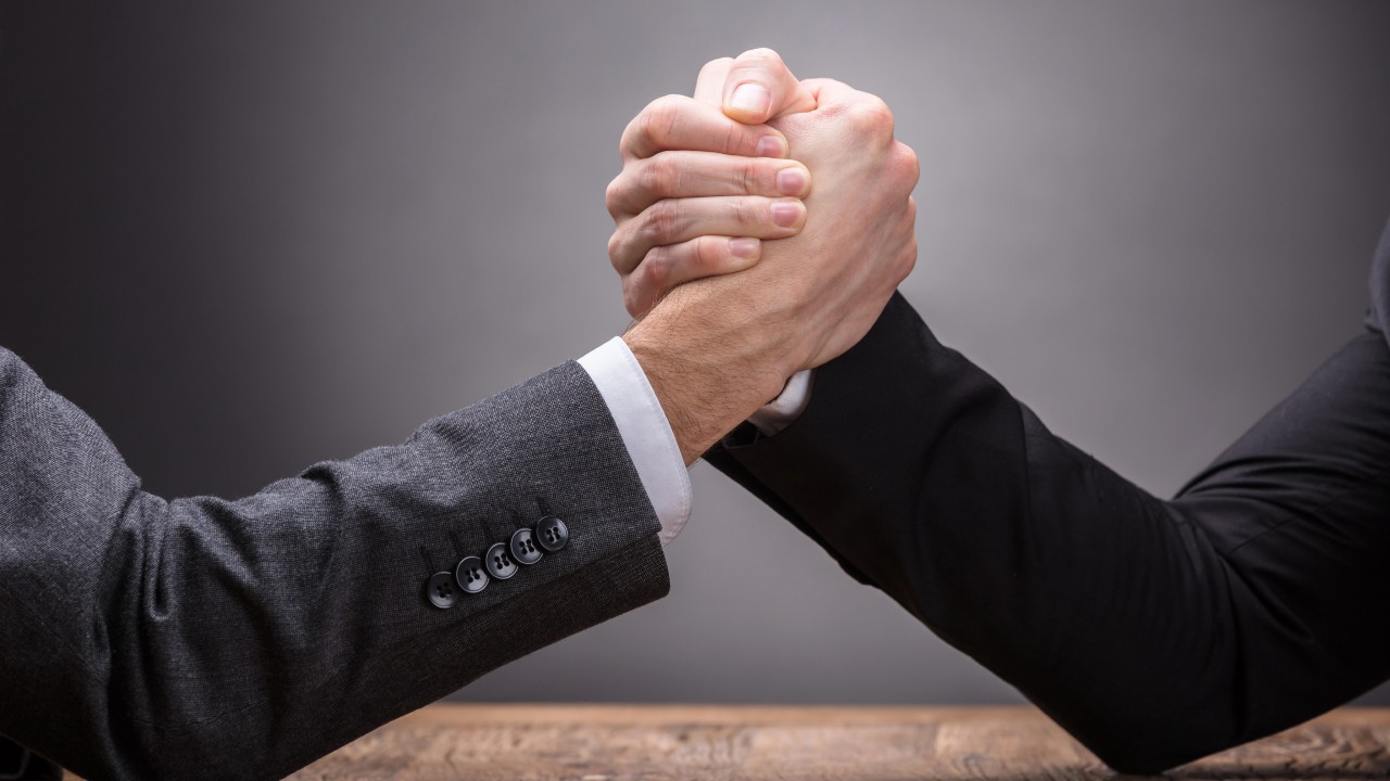 Due persone si sfidano a braccio di ferro su un tavolo di legno, entrambi con blazer scuri, alludendo a un ambiente professionale. Il riferimento va all’arbitro assicurativo che deve risolvere le controversie nelle polizze vita.