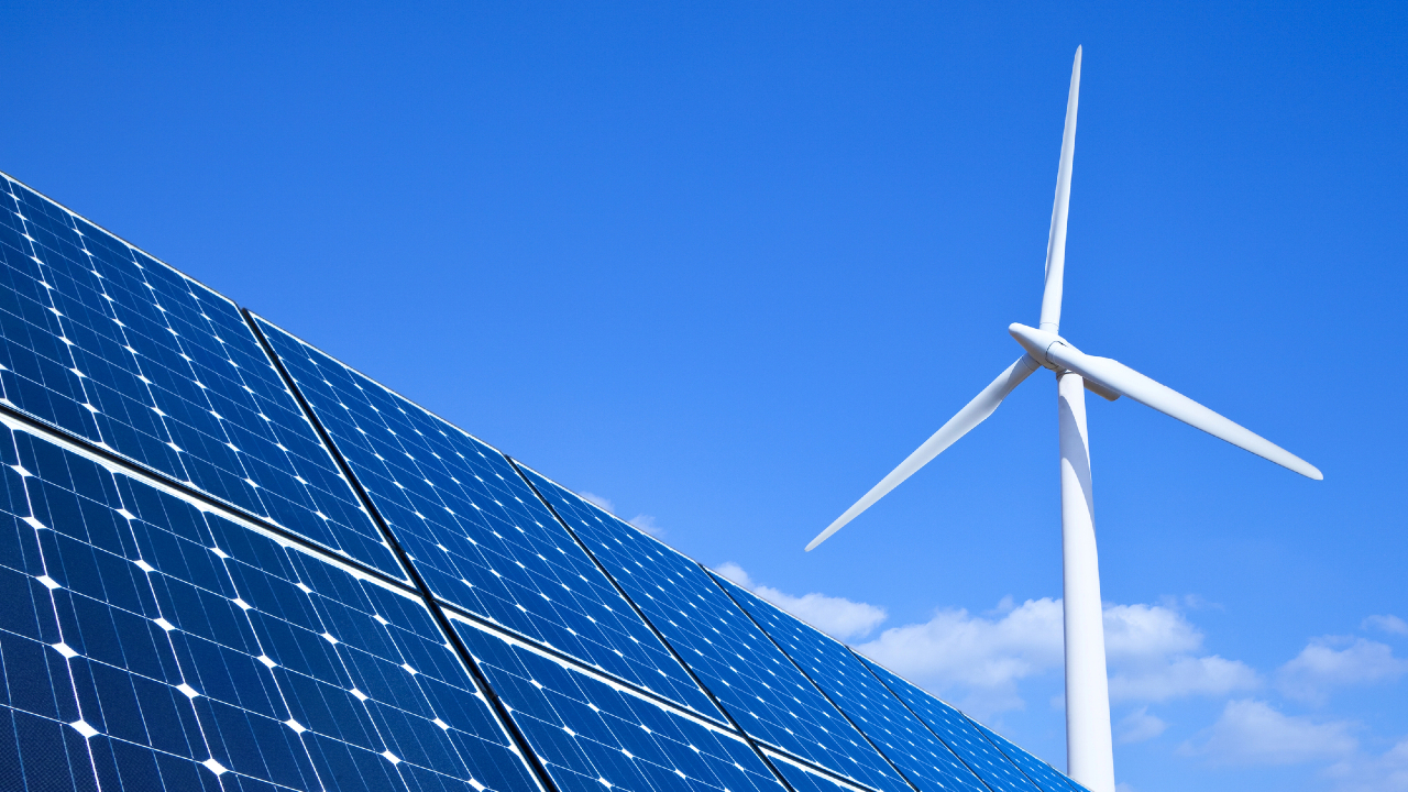 Un pannello solare e una turbina eolica bianca sotto un cielo azzurro e terso, a simboleggiare l'azione verso l'energia rinnovabile. Il pannello solare è angolato perfettamente, mentre l'imponente turbina eolica si erge maestosa sullo sfondo di nuvole sparse.