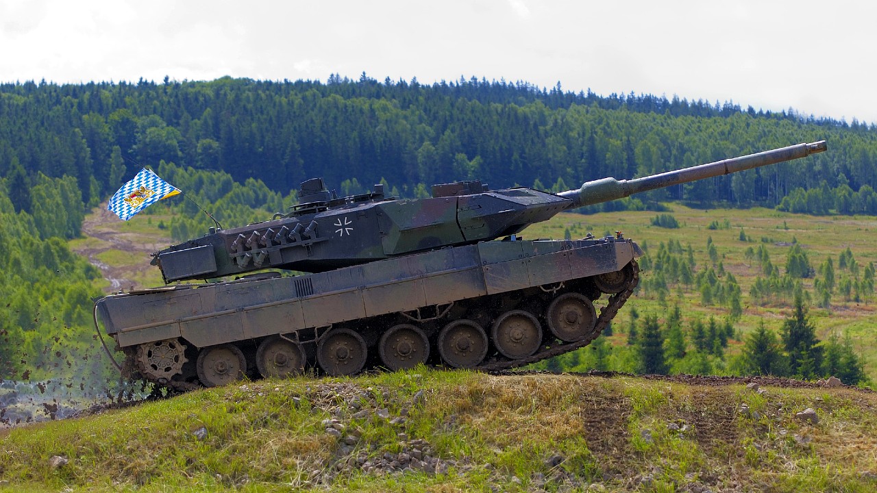Un carro armato militare con un design mimetico sta guidando su una collina erbosa. Una bandiera a scacchi blu e bianca con uno stemma è montata sul carro armato. Sullo sfondo, c'è una fitta foresta sotto un cielo nuvoloso.