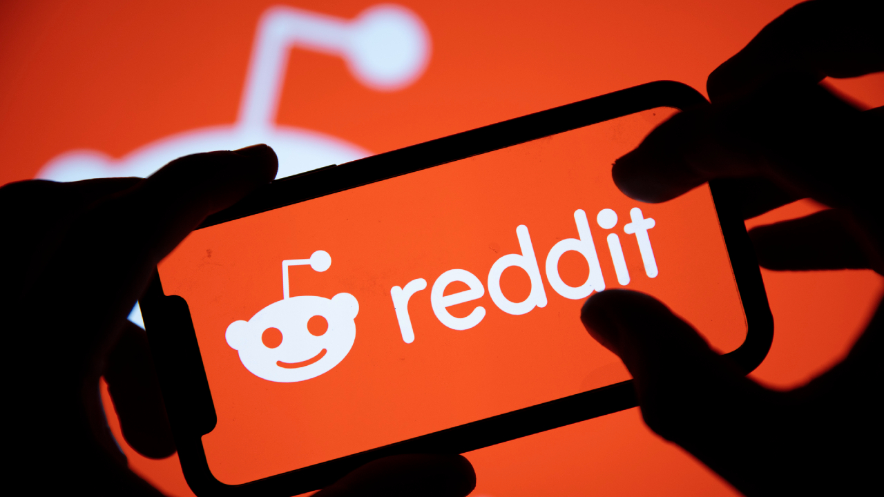 Una persona che tiene in mano uno smartphone con il logo di Reddit sullo sfondo, su un logo Reddit più grande e sfocato. L'immagine, inondata dai colori arancioni e bianchi tipici del brand, rispecchia sottilmente la natura dinamica delle azioni che si svolgono all'interno di vivaci comunità online.