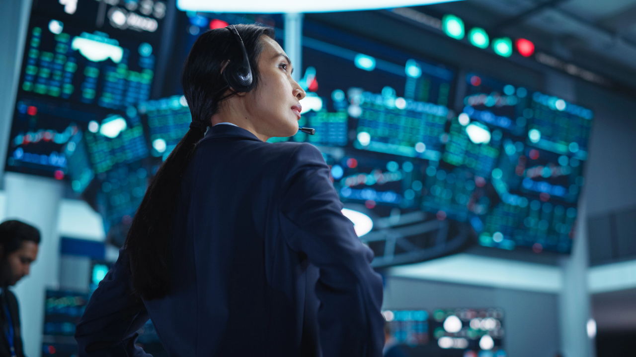 Una persona che indossa un visore sta in piedi con sicurezza in una sala di controllo affollata, piena di grandi schermi che mostrano grafici e dati complessi. L'ambiente suggerisce un monitoraggio ad alta tecnologia o un trading floor di azioni, con più postazioni e attività mirate.