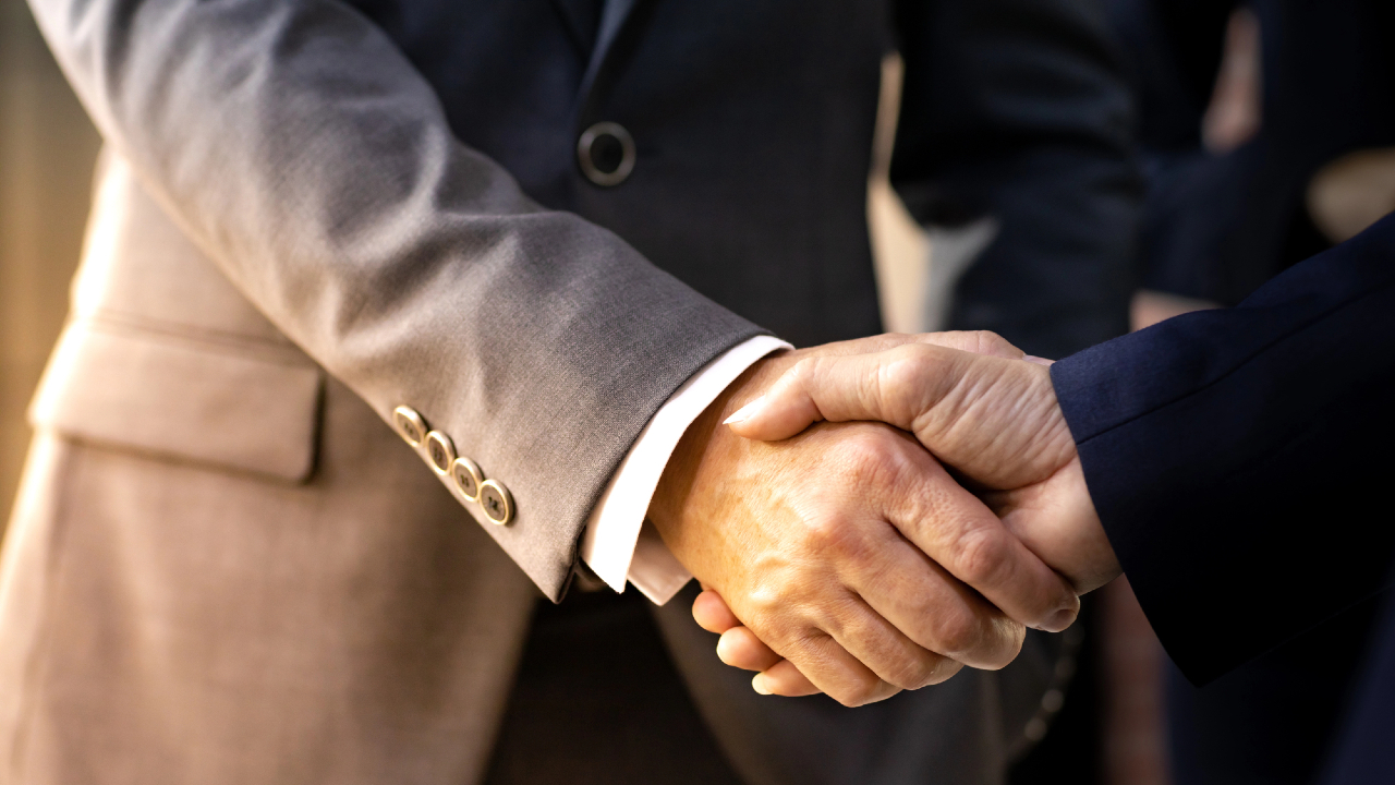 Due individui in abiti da lavoro si stringono la mano, a simboleggiare un accordo o una partnership. Uno indossa un completo grigio, l'altro uno blu scuro. Lo sfondo leggermente sfocato allude sottilmente a raffinatezza, simile a una collaborazione Ifis Euclidea.