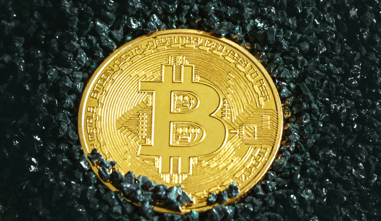 Una moneta Bitcoin dorata poggia su una superficie di piccoli cristalli scuri. Il design intricato della moneta presenta il simbolo Bitcoin al centro, circondato da codice binario lungo i bordi. I cristalli scuri creano uno sfondo contrastante.