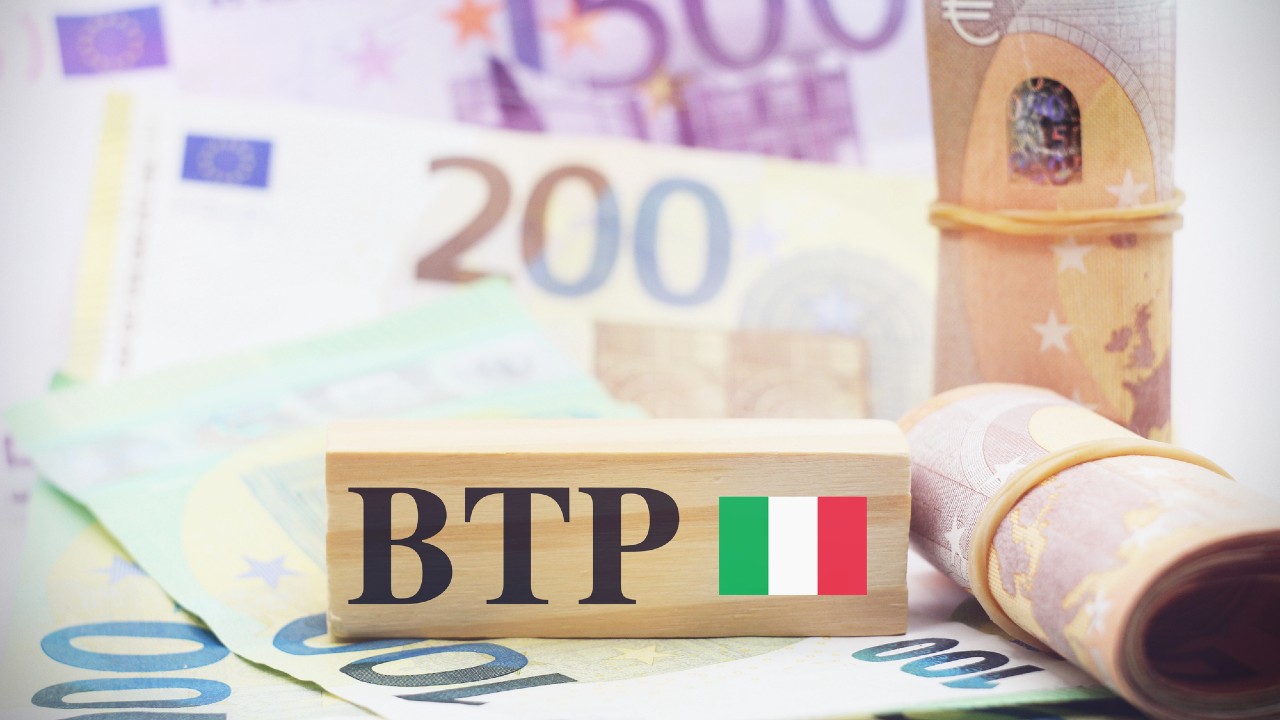 Un blocco di legno con la scritta "BTP" e la bandiera italiana è posto su uno sfondo di varie banconote in euro, tra cui quelle da 500, 200 e 100 euro. È visibile anche un fascio arrotolato di banconote, legato con un elastico.