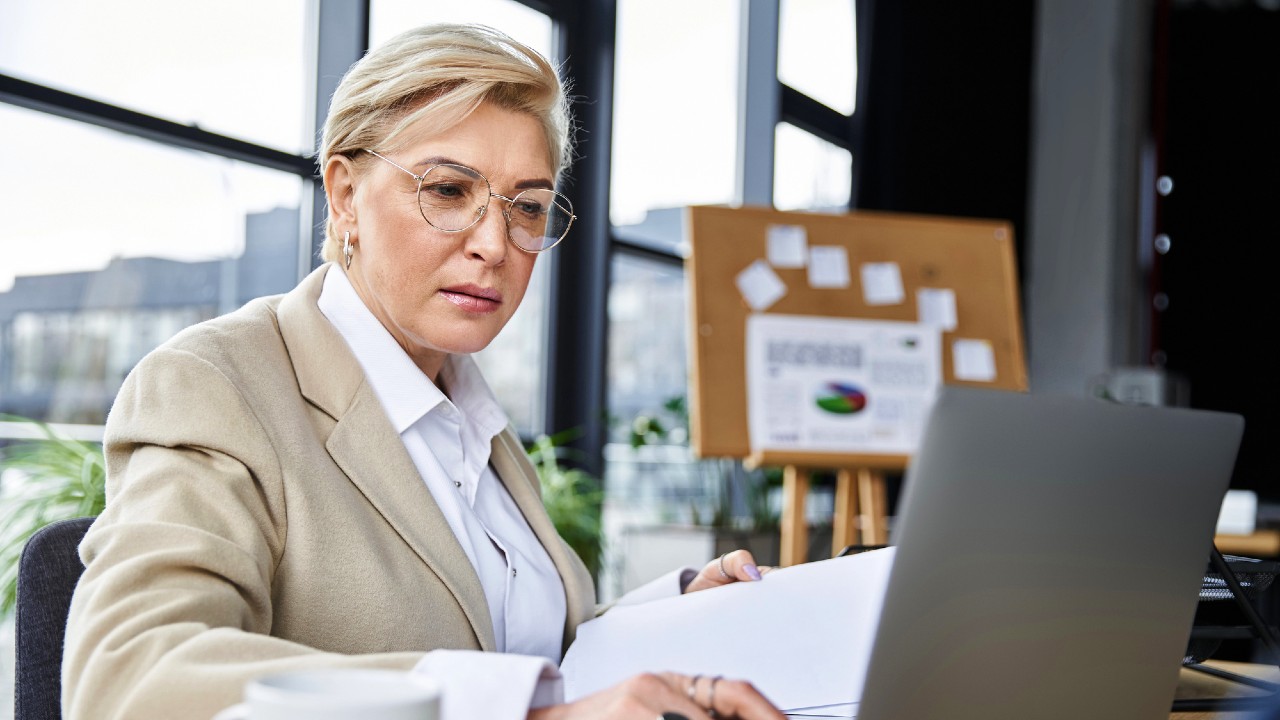 Una persona con i capelli biondi corti e gli occhiali lavora intensamente a un computer portatile in un moderno ufficio. Tiene in mano dei documenti, con una bacheca di sughero e grandi finestre visibili sullo sfondo. Una tazza è appoggiata sulla scrivania accanto a loro.