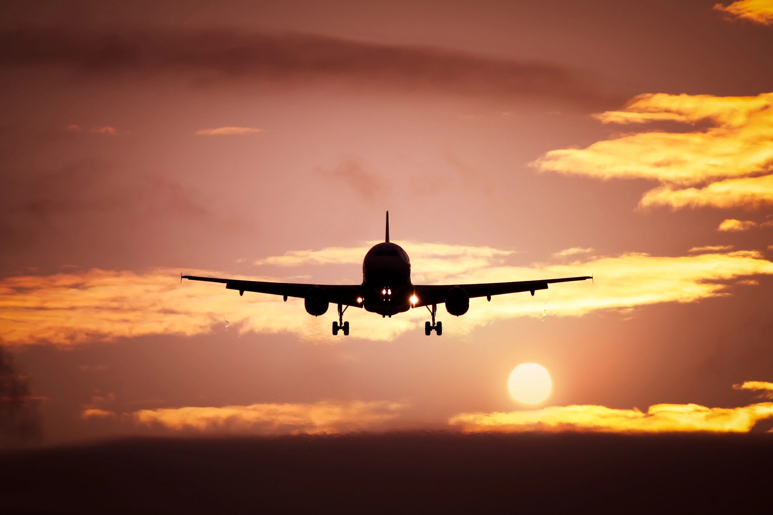 La silhouette di un aereo in atterraggio è sullo sfondo di un cielo al tramonto spettacolare, che ricorda un certificato del settore aeronautico, con il sole parzialmente visibile sotto. Il cielo è pieno di nuvole dorate, creando un'atmosfera calda e serena.