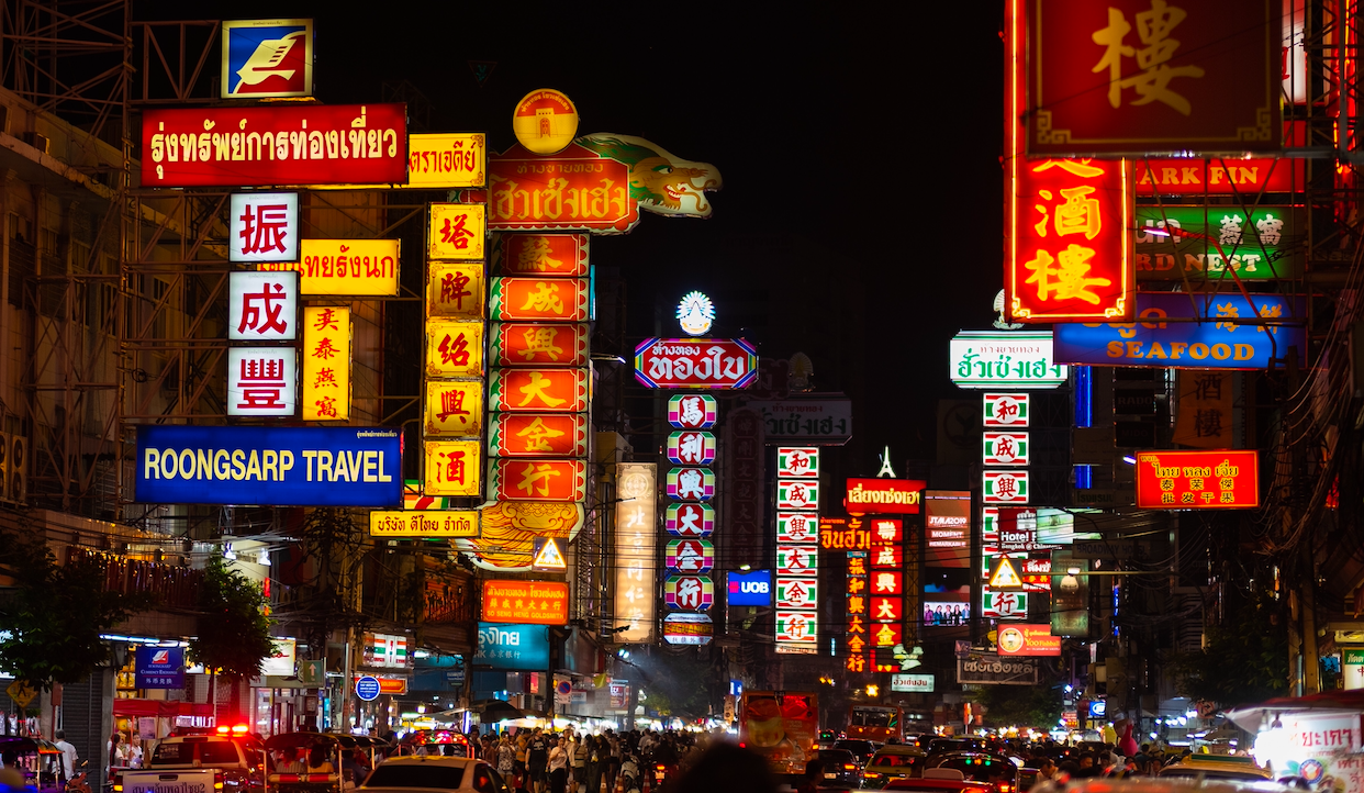 Una scena di strada notturna e movimentata in una città, illuminata da numerose insegne al neon colorate con scritte asiatiche. Le insegne vivaci creano un'atmosfera vivace, che si riflette sulla strada trafficata piena di persone e veicoli.