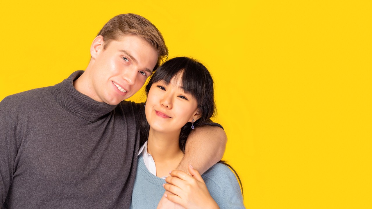 Un esempio di coppie internazionali sorridenti posa su uno sfondo giallo brillante. L'uomo, con un dolcevita grigio, ha il braccio intorno alle spalle della donna. La donna, che indossa un maglione azzurro, si avvicina.
