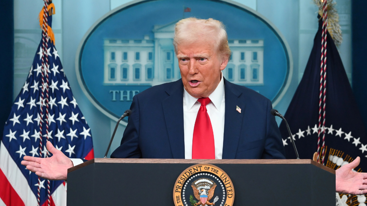 Un uomo in giacca e cravatta rossa gesticola dinamicamente mentre parla su un podio con il sigillo presidenziale degli Stati Uniti, discutendo di azioni cruciali sulle politiche. Bandiere americane e uno sfondo che ricorda la Casa Bianca esaltano la sua autorevole presenza.