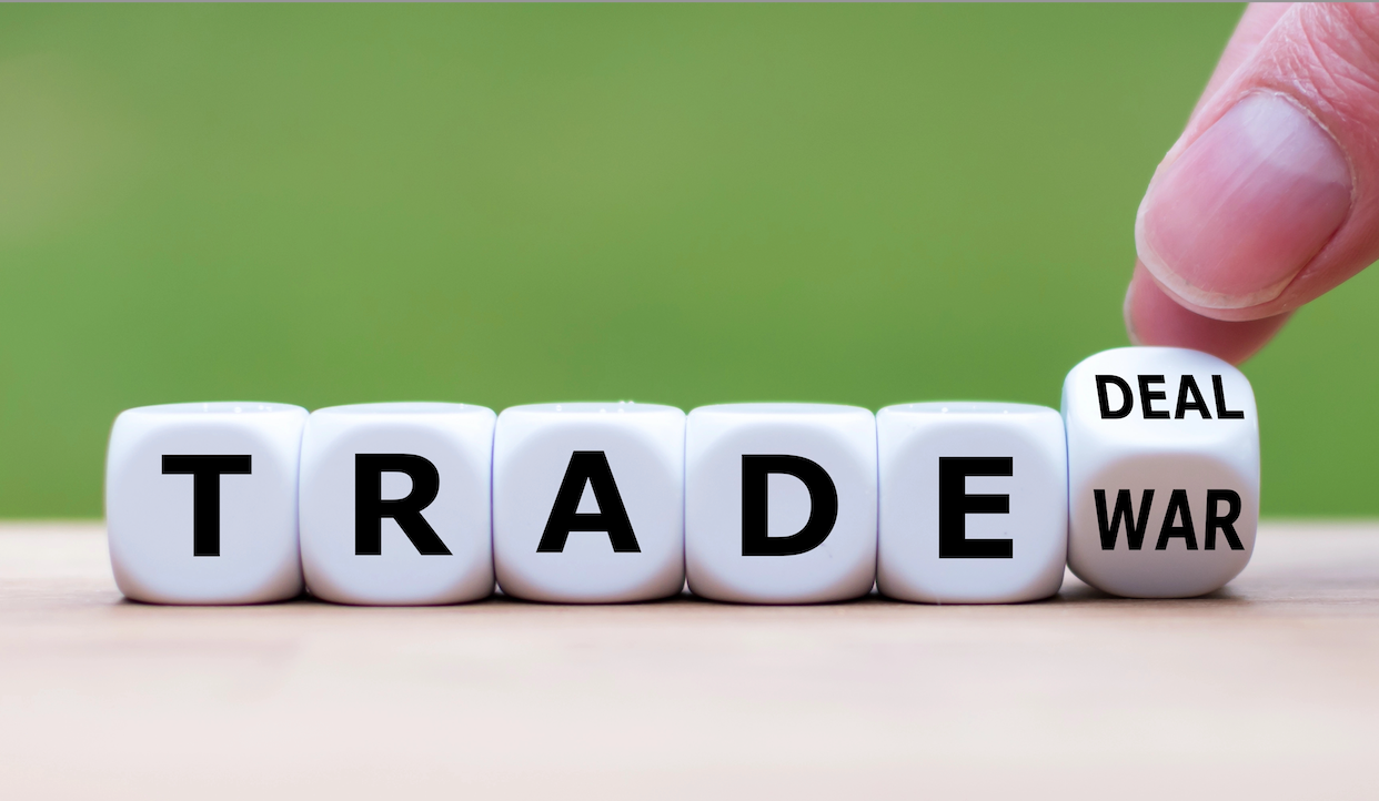 Primo piano di sei cubi bianchi su una superficie di legno, che compongono la scritta "TRADE". Una mano gira l'ultimo cubo che mostra "DEAL", che ha anche "WAR" sul lato opposto. Lo sfondo è di un verde sfocato, simile all'erba.