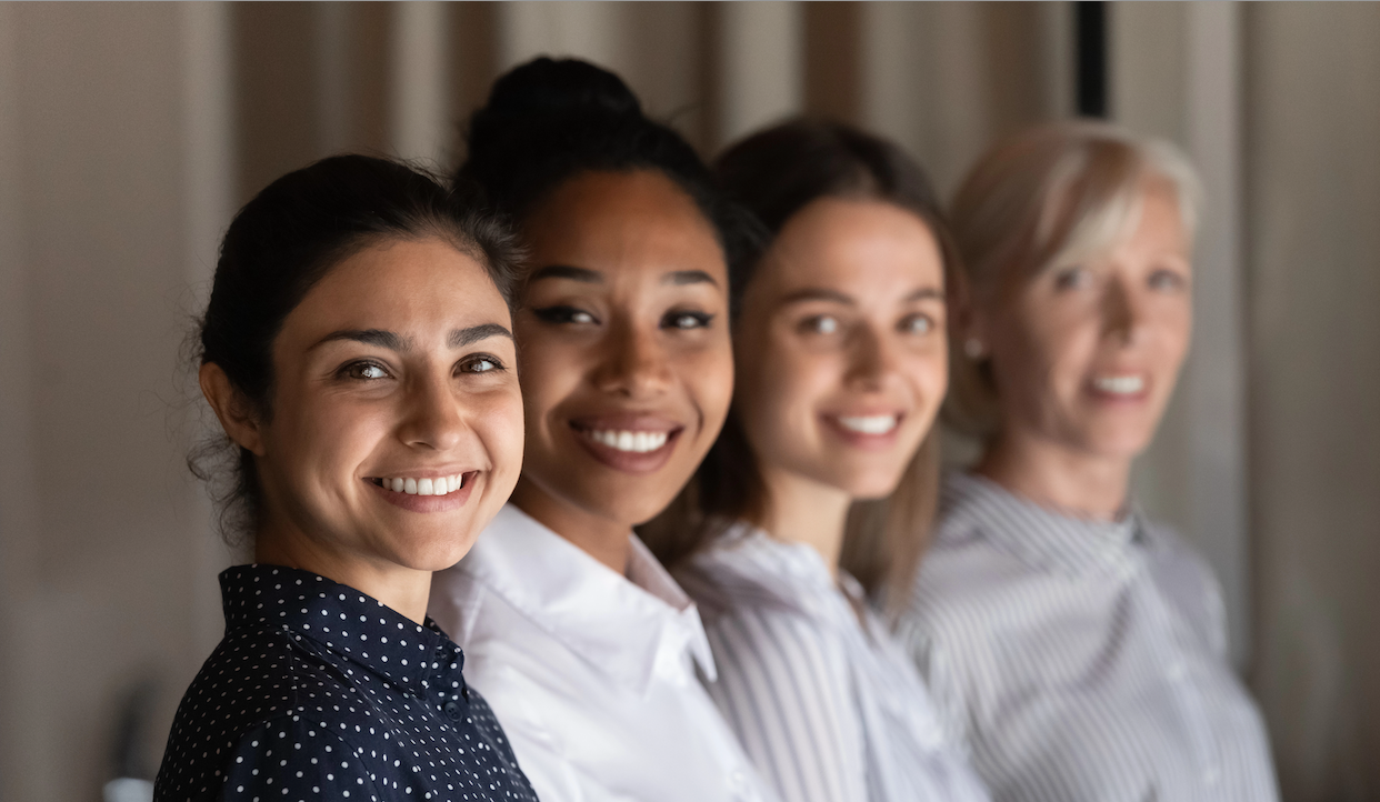 Quattro donne in fila, sorridenti alla telecamera. Sono vestite in abiti casual eleganti e variano per età e aspetto. Il gruppo è al chiuso e lo sfondo è leggermente sfocato.
