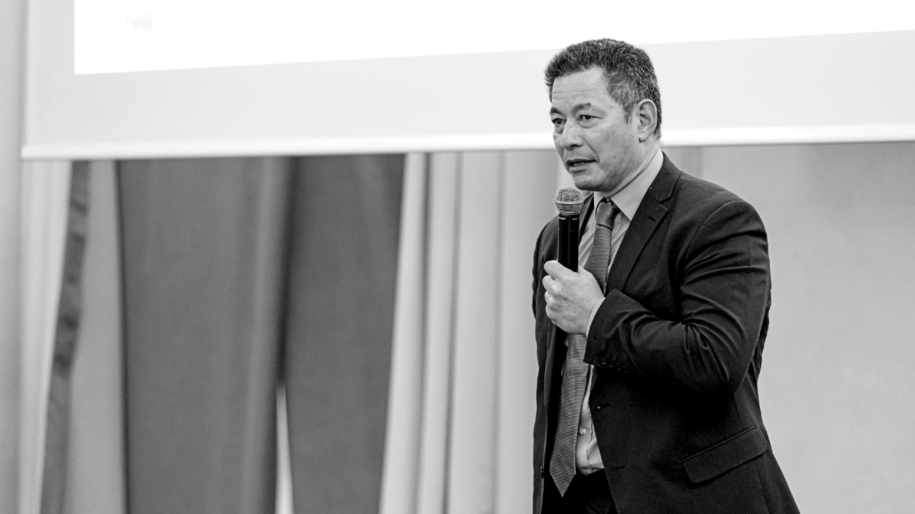 Un uomo in giacca e cravatta parla in un microfono, in piedi davanti a un grande schermo in una sala conferenze. L'immagine, resa in bianco e nero, lo cattura in un ambiente formale, probabilmente mentre tiene una presentazione o un discorso.