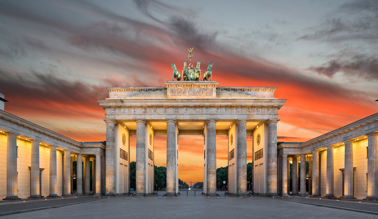 La Porta di Brandeburgo a Berlino illuminata al tramonto, con un cielo spettacolare di tonalità arancioni e rosse. Il monumento neoclassico presenta colonne e una statua di carro in cima, creando una silhouette sorprendente contro il cielo colorato.