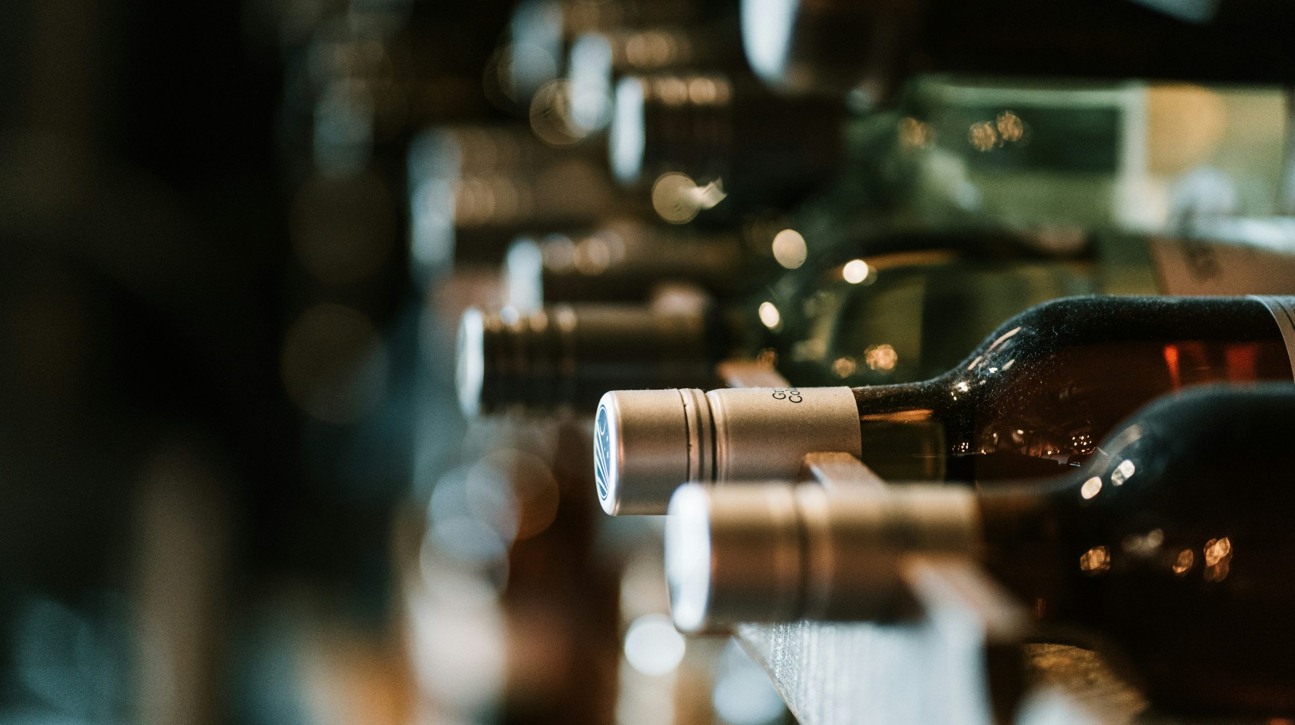 Primo piano di bottiglie di vino disposte orizzontalmente su una rastrelliera di legno, che mostrano varie etichette e tappi. Lo sfondo è leggermente sfocato, focalizzato sulla fila di bottiglie, evocando un senso di cantina rustica e caldamente illuminata.