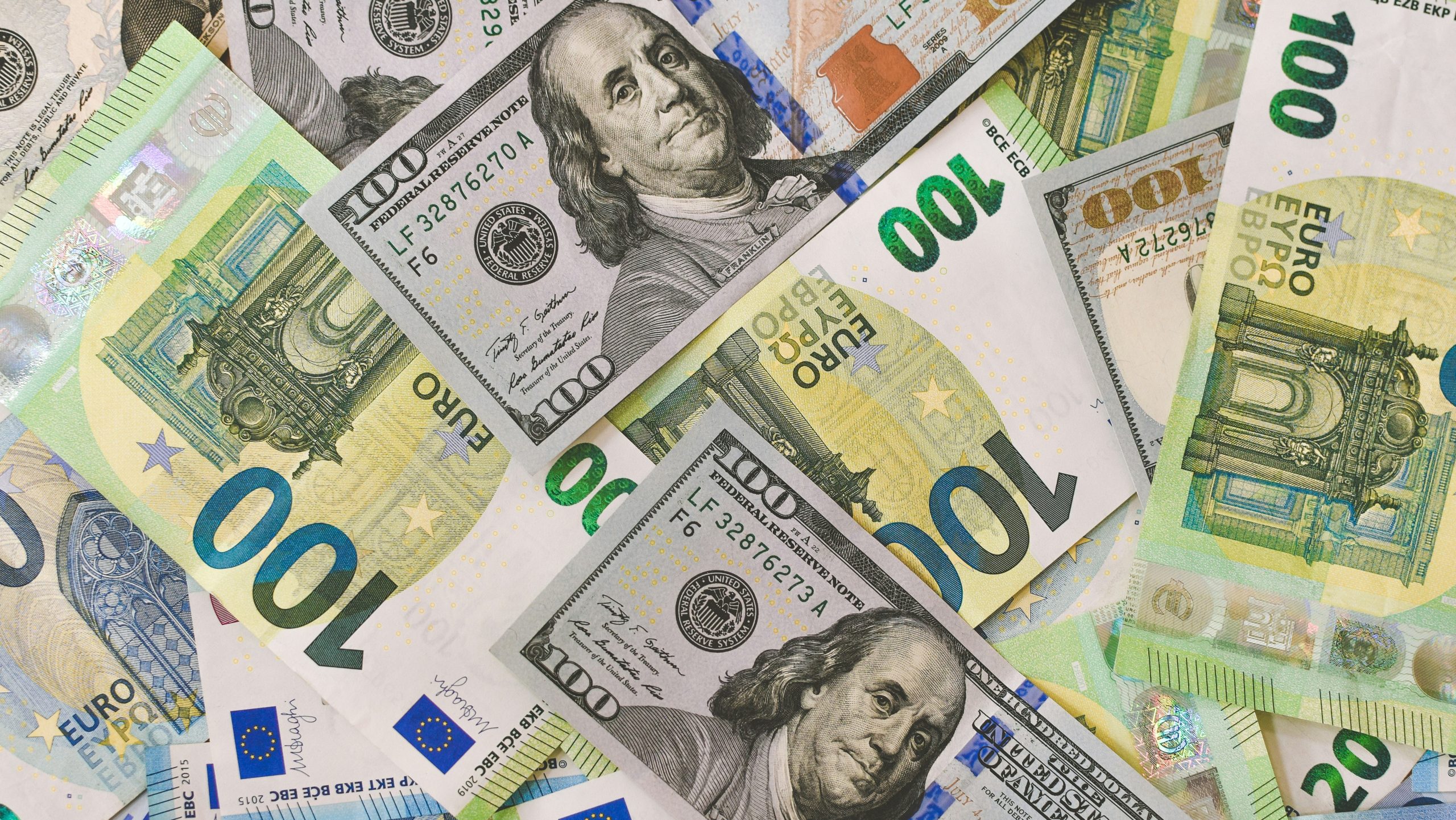 Una pila sparsa di banconote assortite, tra cui banconote da 100 dollari statunitensi raffiguranti Benjamin Franklin e banconote da 100 euro europee, giace accanto a una brochure sui fondi obbligazionari Carmignac. Le banconote si sovrappongono, mostrando colori e design diversi.