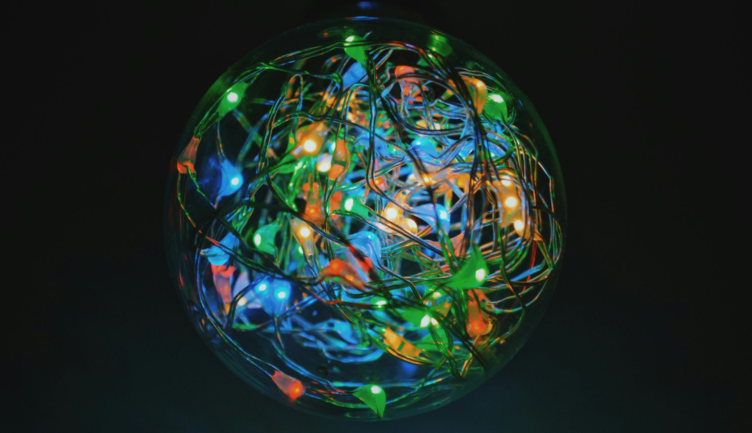 Una sfera di vetro trasparente riempita con fili intrecciati di luci LED colorate: verde, blu, arancione e giallo. Le luci brillano intensamente su uno sfondo scuro, creando un aspetto vibrante e festoso.