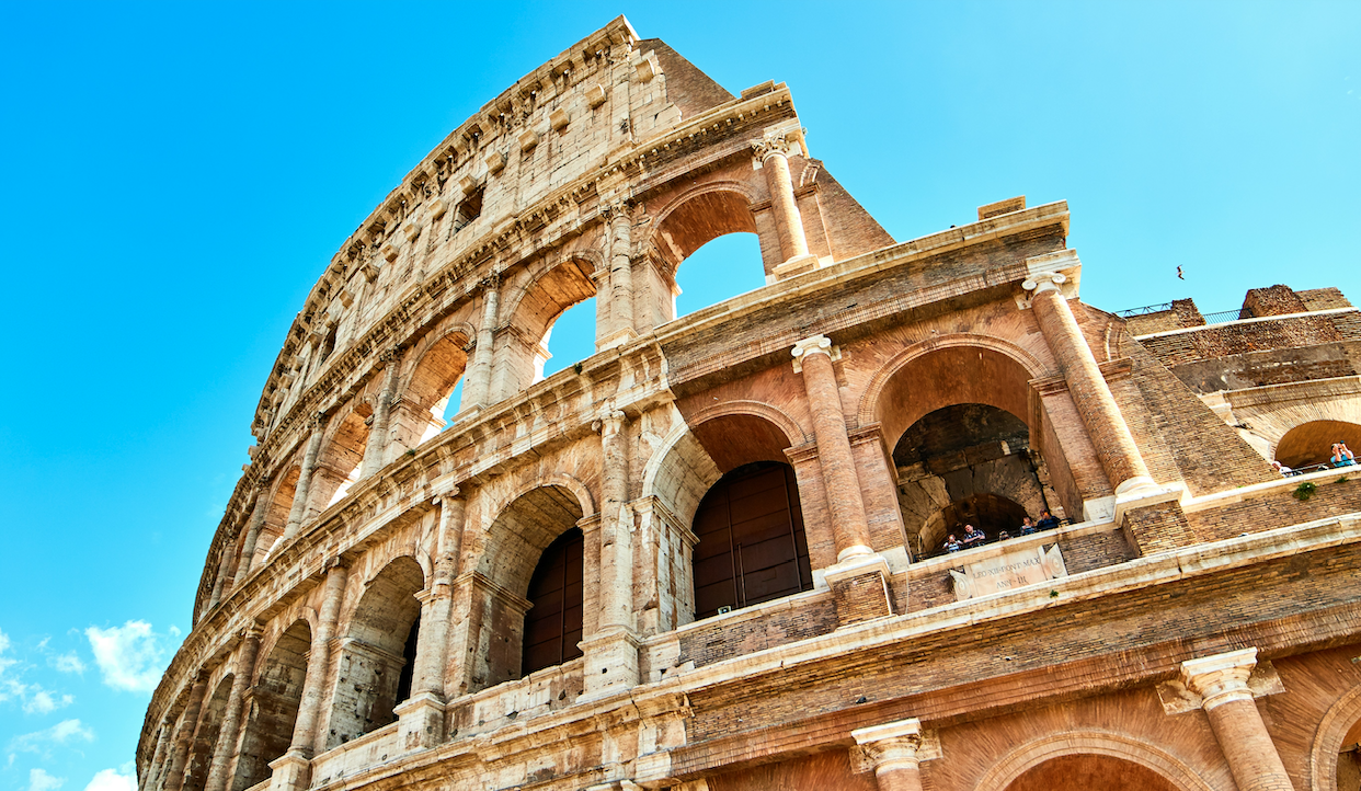 L'immagine mostra una vista angolata del Colosseo contro un cielo azzurro terso. Gli archi dell'antico anfiteatro romano e la facciata in pietra erosa sono ben visibili, evidenziandone l'architettura storica.