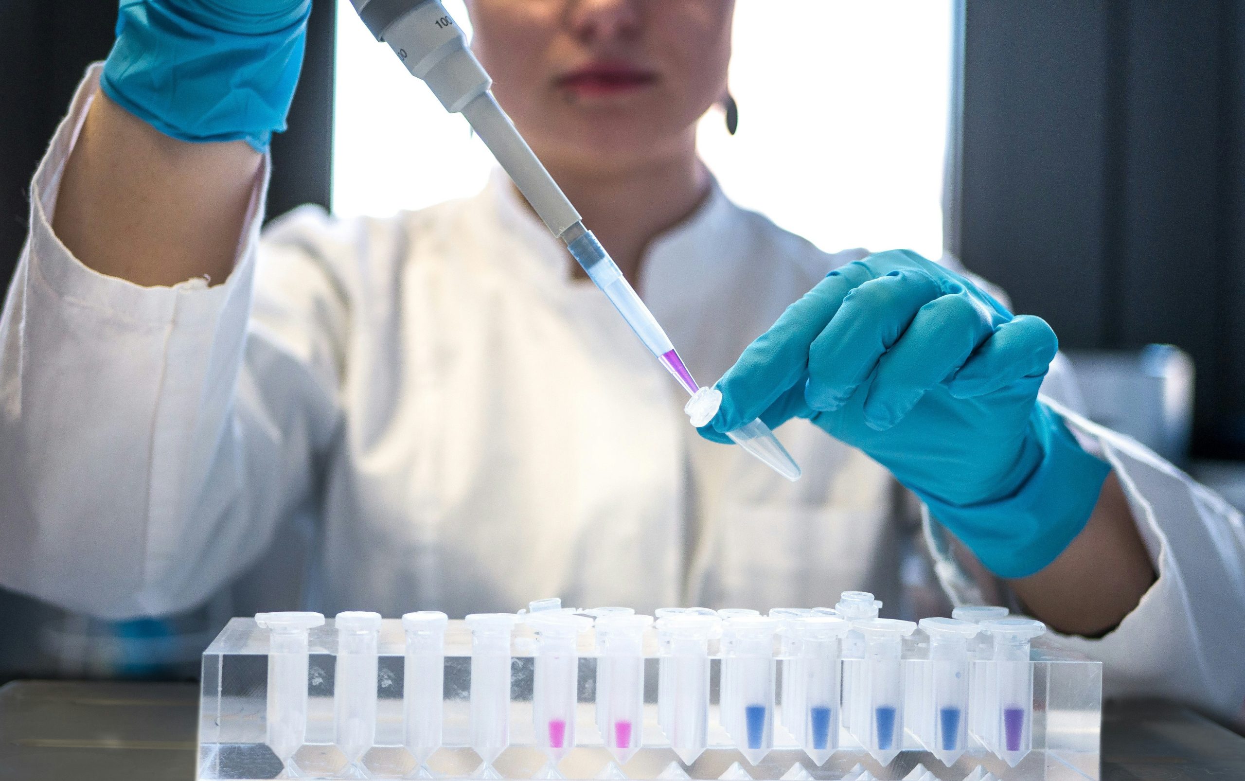 Uno scienziato in camice bianco e guanti blu usa con attenzione una pipetta per trasferire il liquido in piccole provette. In primo piano, diverse provette, alcune contenenti liquidi colorati, sono organizzate in un supporto, esemplificando i progressi nella ricerca sanitaria relativa al GLP-1.