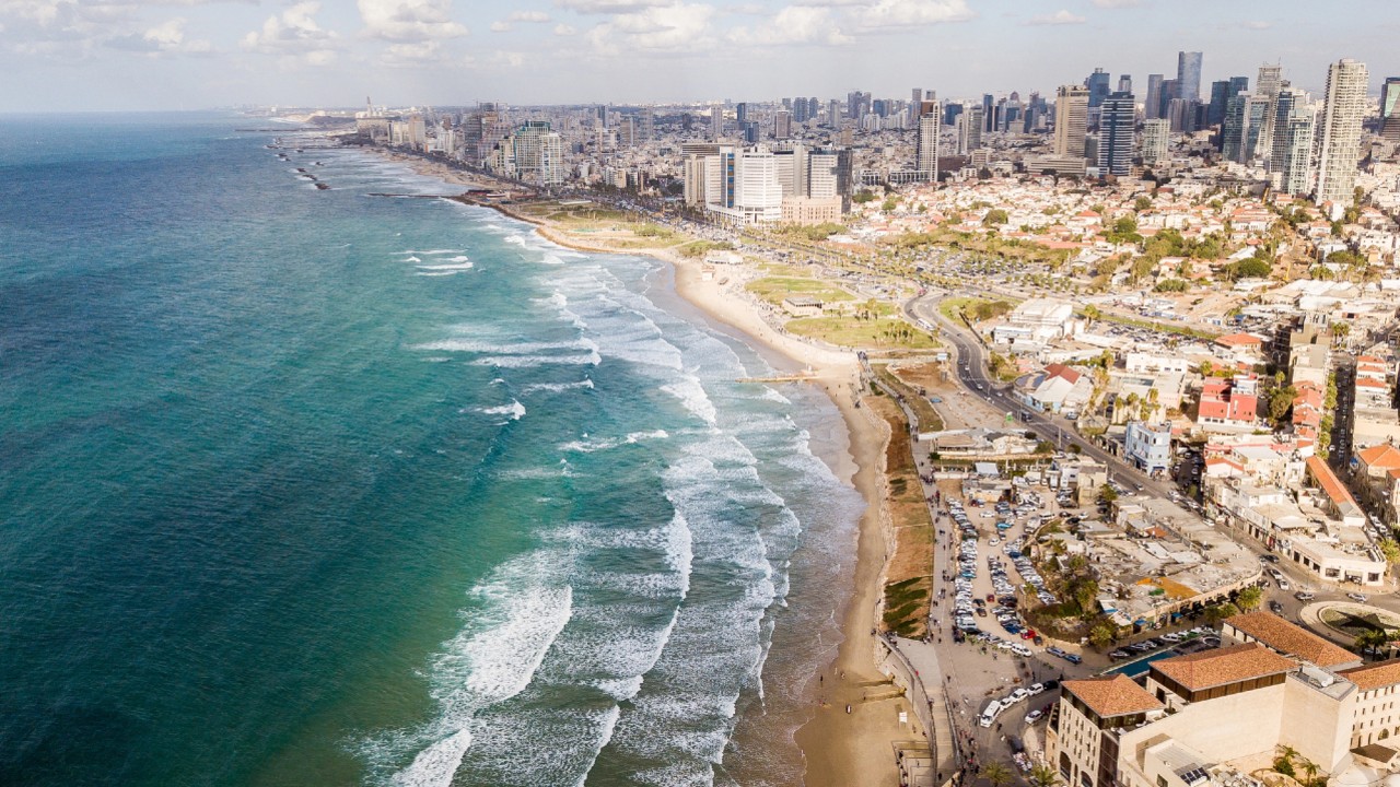 Vista aerea di una città costiera con alti edifici e aree urbane sulla destra e una spiaggia sabbiosa con onde sulla sinistra. La costa curva dolcemente, con un mix di moderni grattacieli e piccoli edifici residenziali sotto un cielo parzialmente nuvoloso.
