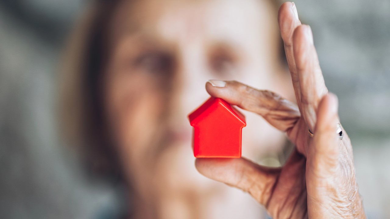 Una persona anziana tiene in mano una piccola casa giocattolo rossa a fuoco, a simboleggiare la vendita della nuda proprietà. Lo sfondo è sfocato, attirando l'attenzione sulla casa