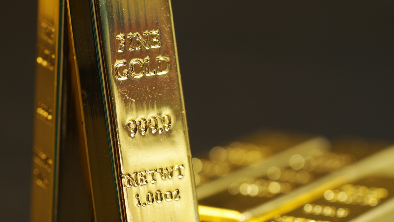 Primo piano di una barra d'oro scintillante contrassegnata con "FINE GOLD 999.9" e "NET WT 1.000 OZ". Sullo sfondo sono presenti altre barre d'oro sfocate, creando un'atmosfera calda e lussuosa.