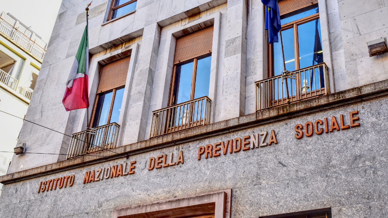 La facciata dell'edificio italiano con una finitura in pietra che mostra "Istituto Nazionale Della Previdenza Sociale" si erge orgogliosa, con diverse finestre e la bandiera italiana. Con un focus sul "pagamento pensioni", si trova sotto un cielo limpido, a simboleggiare la sicurezza per i pensionati.