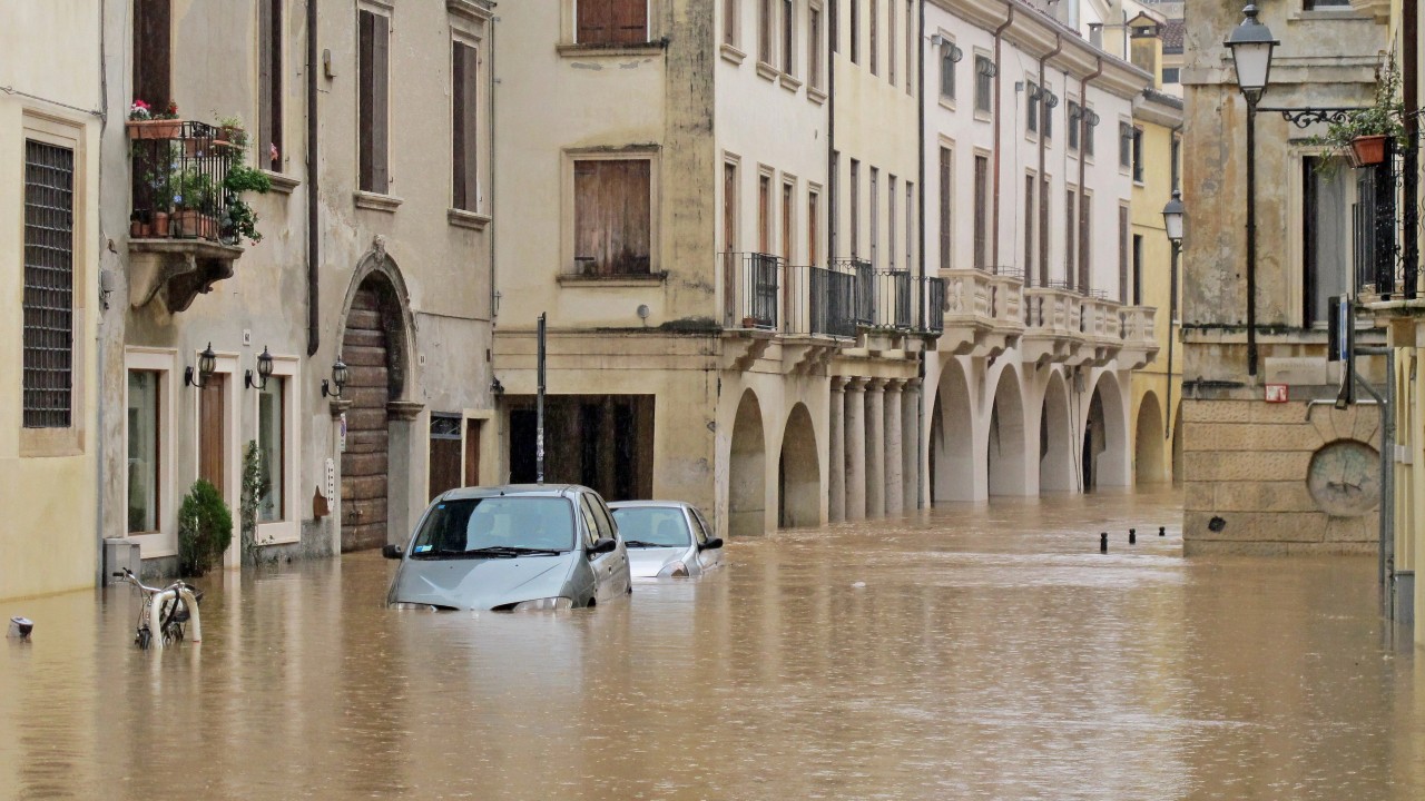 Una strada allagata con auto e biciclette sommerse, acqua a metà dei veicoli, case ed edifici. Il riferimento va alla necessità di stipulare per le imprese una polizza catastrofale, obbligatoria nel 2025.