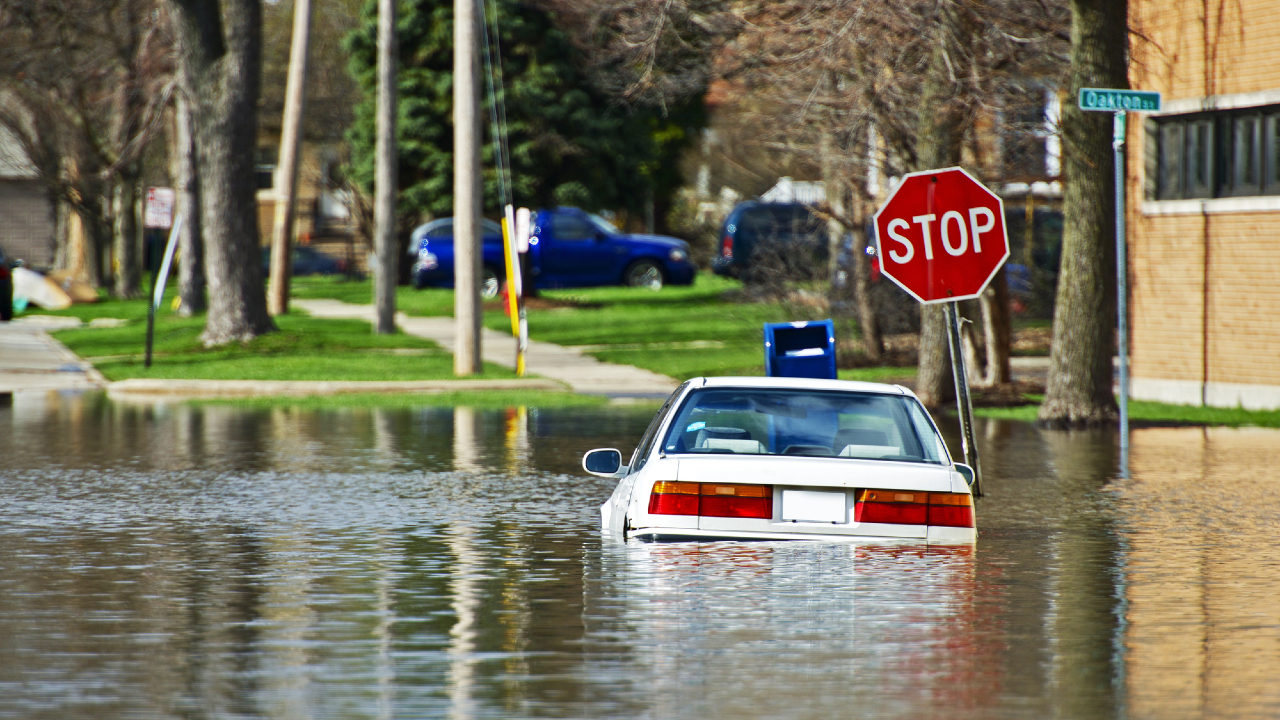 Un'auto bianca è parzialmente sommersa dall'acqua in una strada residenziale, evidenziando la necessità di una polizza catastrofale. Un segnale di stop si trova lì vicino, con alberi e case che costeggiano la strada sotto un cielo coperto.