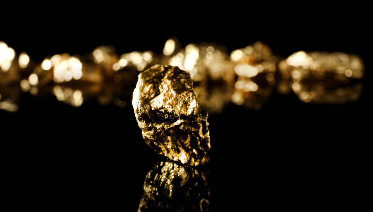 Primo piano di una pepita d'oro lucente su una superficie nera riflettente, con diverse pepite d'oro sfocate sullo sfondo. L'illuminazione mette in risalto le texture e la lucentezza della pepita.