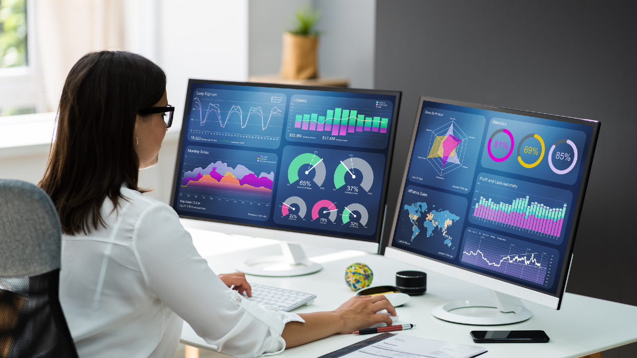 Una persona è seduta a una scrivania e guarda due monitor di computer che mostrano visualizzazioni di dati colorate, tra cui grafici, diagrammi e mappe. L'ambiente dell'ufficio vanta una finestra luminosa sullo sfondo, creando un'atmosfera serena perfetta per un'analisi meticolosa prima della pianificazione della pensione.