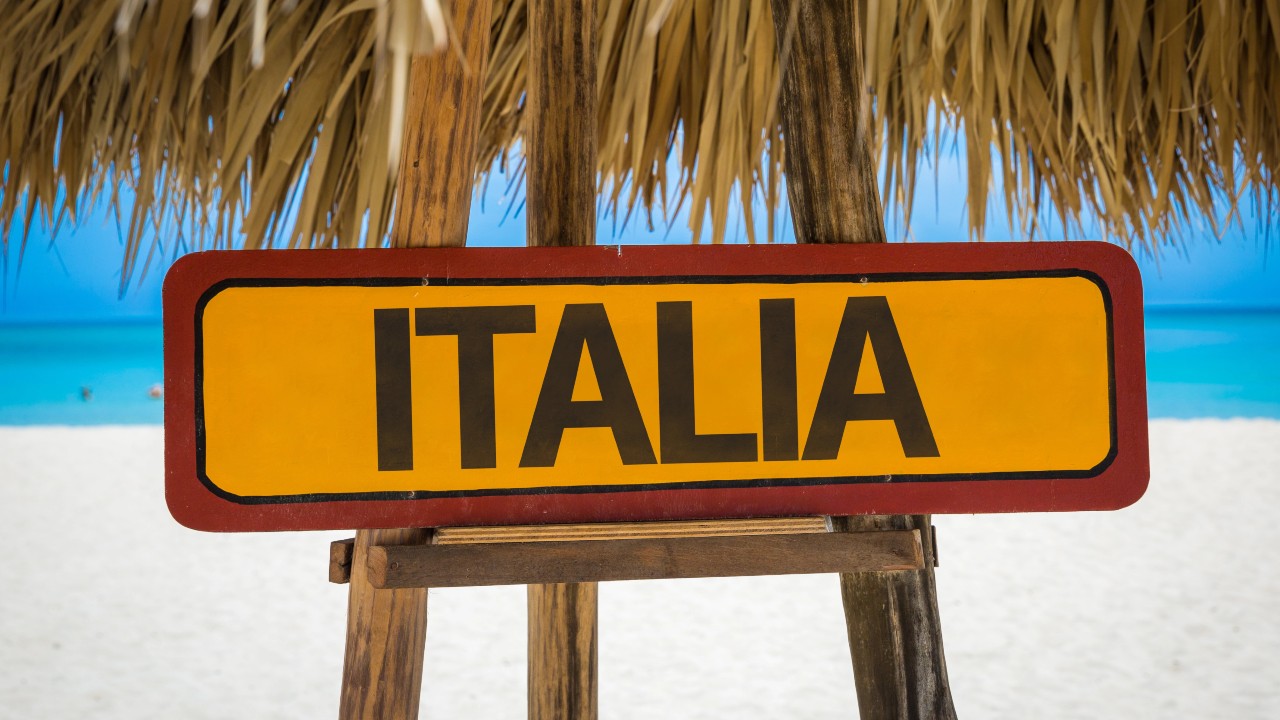 Un cartello giallo con la parola "Italia” in grassetto nero è esposto su un cavalletto di legno sotto un tetto di paglia. Sullo sfondo, una spiaggia di sabbia bianca e un oceano azzurro e limpido sotto un cielo luminoso a indicare l’Italia come un paradiso fiscale legato al tema delle agevolazioni fiscali per lavoratori in caso di rientro in Italia dall’estero.