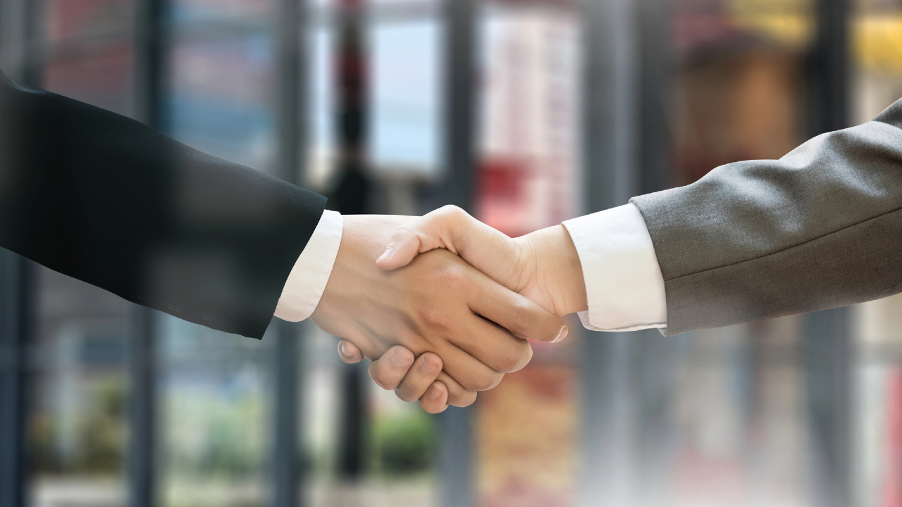 Due persone in abiti da lavoro si stringono la mano; una indossa un completo nero, l'altra un completo grigio. Lo sfondo è sfocato, con grandi finestre tipiche degli ambienti delle banche, che suggeriscono un'atmosfera professionale.