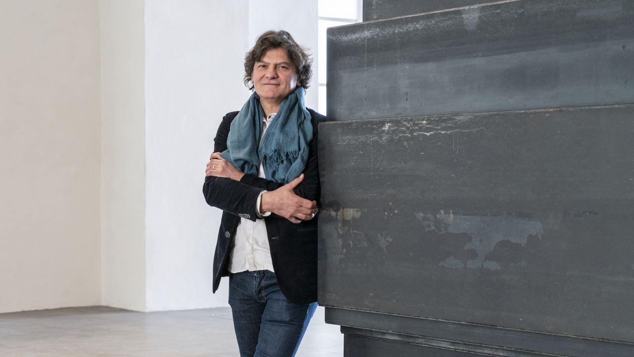 Una persona con i capelli mossi e una sciarpa è in piedi con le braccia incrociate, appoggiata a una grande struttura metallica scura in una stanza minimalista. Indossa un blazer nero e jeans, con la luce naturale proveniente da una finestra che illumina la scena.