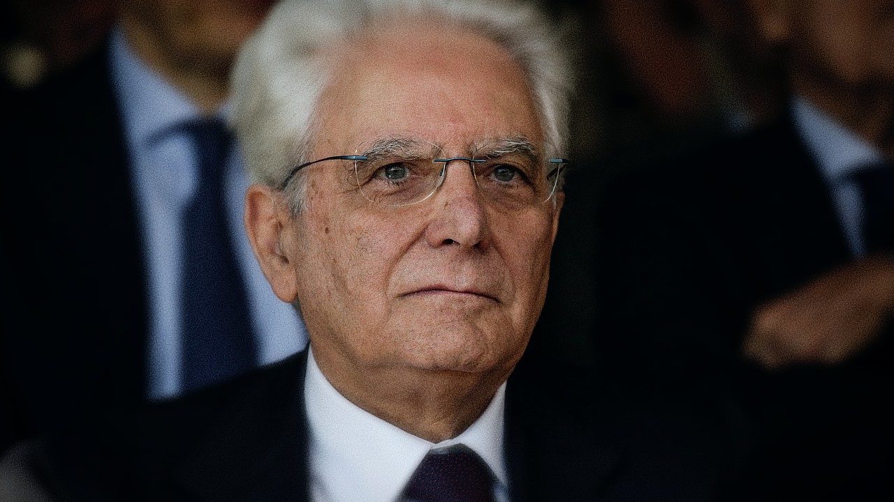Un uomo anziano con i capelli bianchi e gli occhiali indossa un completo e una cravatta. Guarda leggermente verso l'alto con un'espressione seria. Lo sfondo è sfocato e scuro.