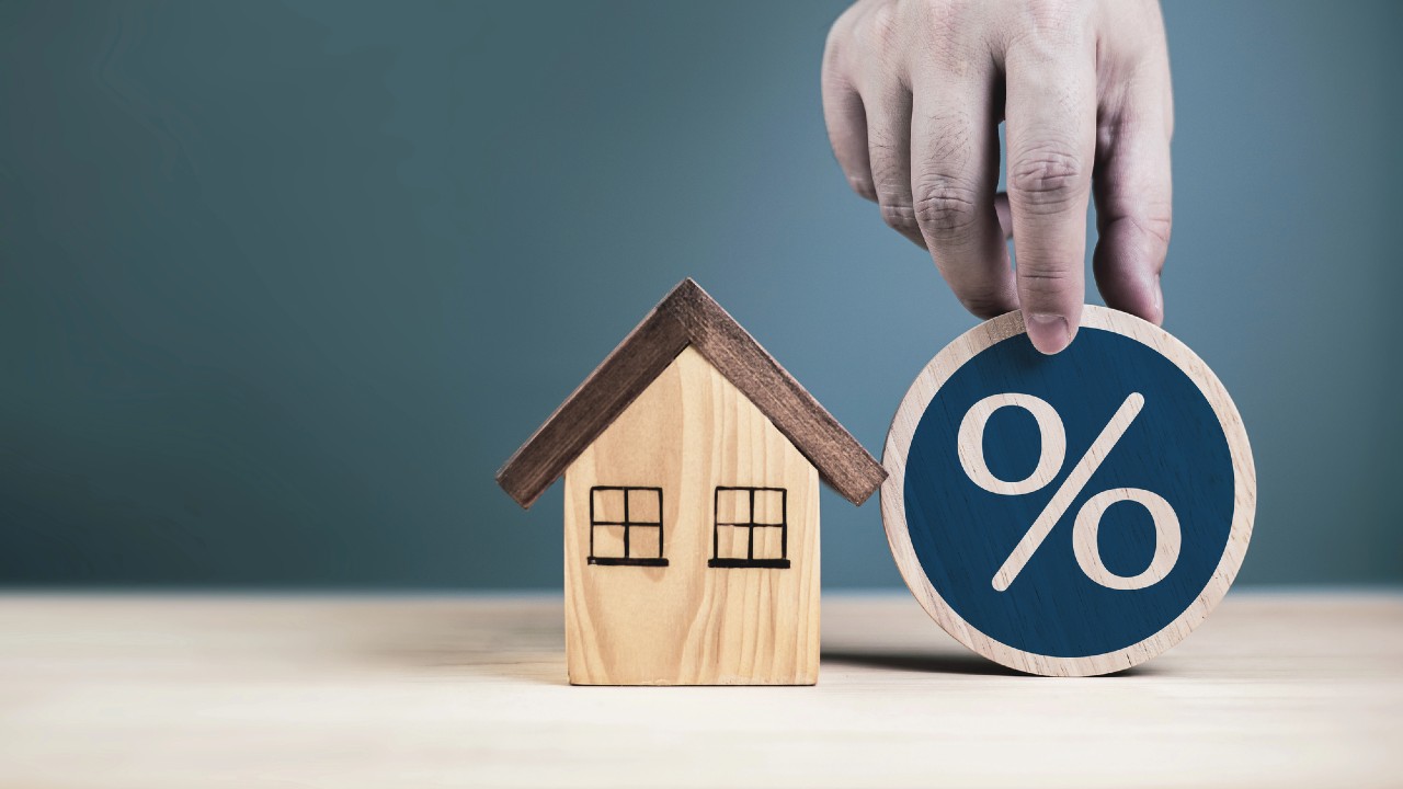 Un modello di casa in legno accanto a una mano che tiene un disco di legno con il simbolo della percentuale, posto su una superficie di legno chiaro su uno sfondo blu.