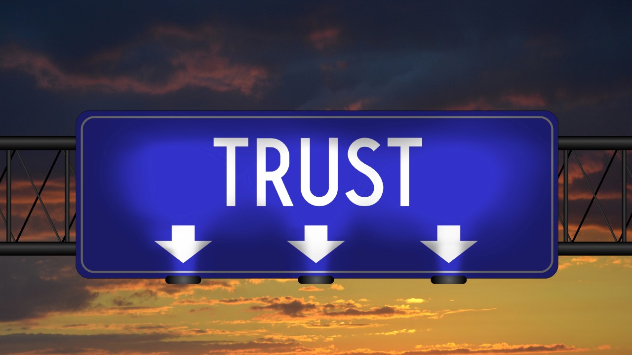 Un cartello rettangolare blu riporta in grassetto la scritta "Trust” in lettere bianche maiuscole, con tre frecce bianche rivolte verso il basso sotto di esso, a indicare il trust come strumento di protezione del patrimonio e della famiglia