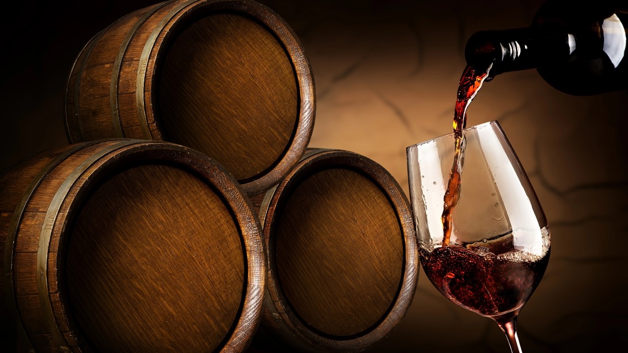 Un vino rosso viene versato elegantemente in un bicchiere da una bottiglia con etichetta artistica sulla destra. Tre botti di legno sono impilate sulla sinistra su uno sfondo caldo e strutturato, creando un'atmosfera rustica e invitante. La foto rimanda a un esempio dove si uniscono vino, arte e impresa