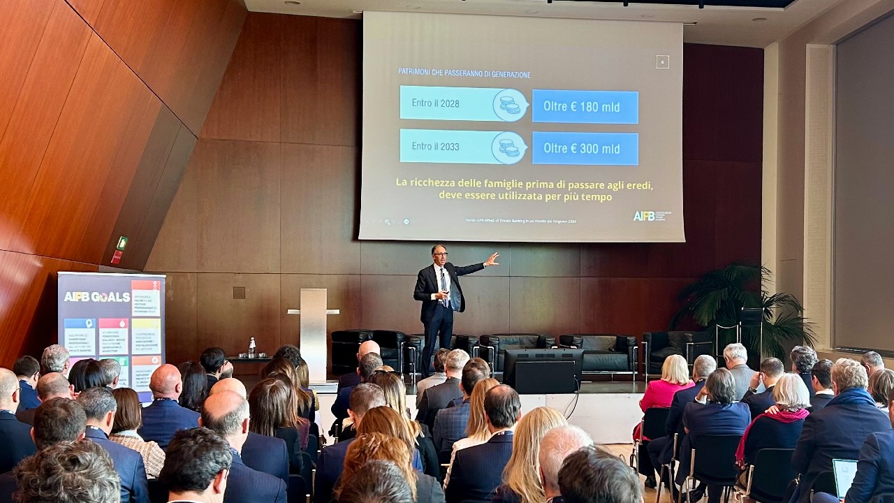 Un uomo in giacca e cravatta sta parlando a un pubblico seduto in una sala conferenze. Un grande schermo dietro di lui mostra informazioni finanziarie e si vede un poster con la scritta "AIBA GOALS".