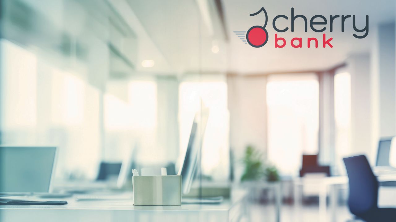 Ufficio moderno e luminoso con pareti in vetro, postazioni di lavoro con computer e una scatola di fazzoletti sulla scrivania. Il logo "Cherry Bank Gestione" con la grafica di una ciliegia è ben visibile sulla parete.