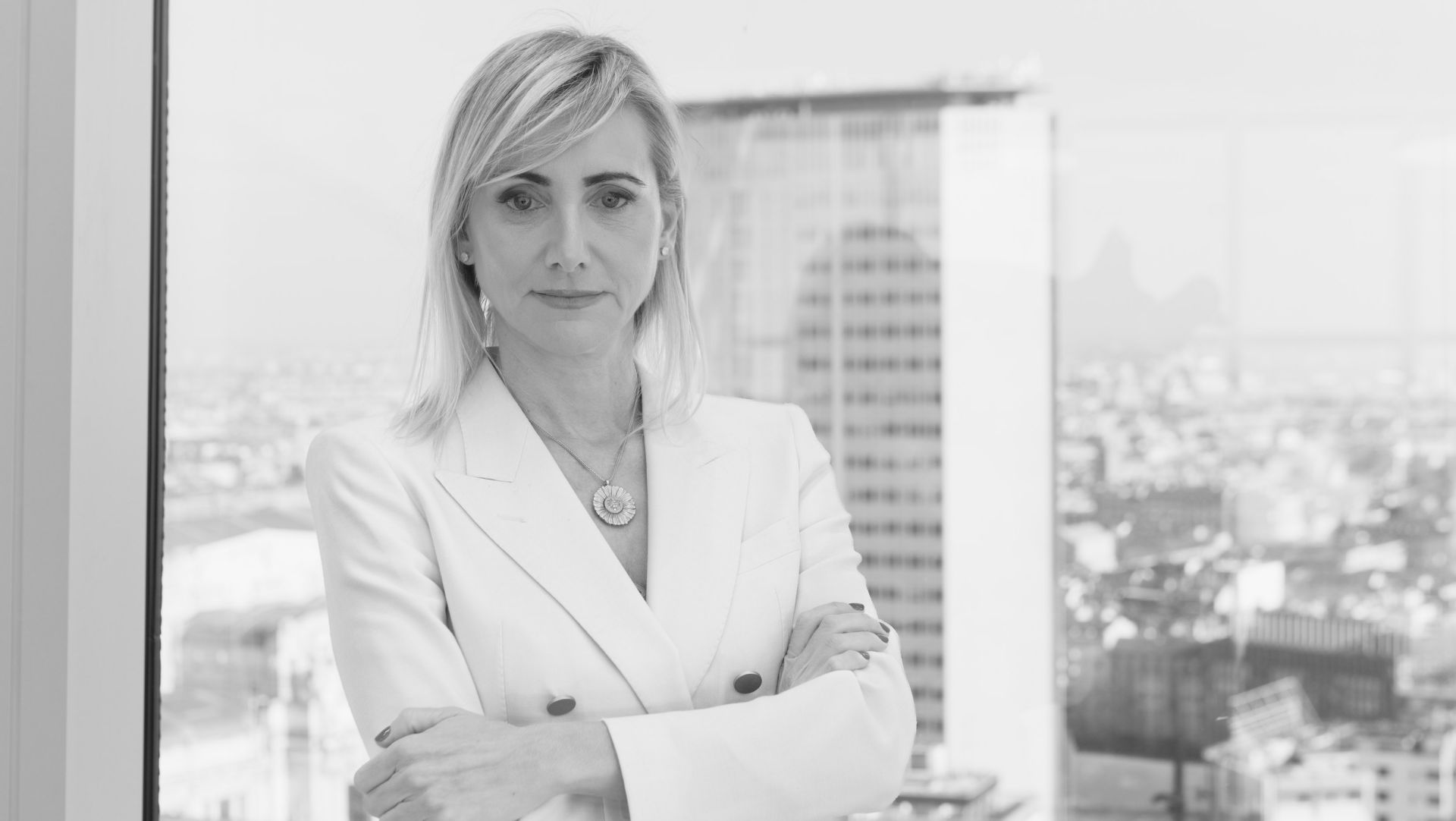 Una persona con i capelli chiari che indossa un elegante abito bianco è in piedi con le braccia incrociate davanti a una finestra. Fuori, il paesaggio urbano sfocato presenta un alto edificio che ricorda l'architettura moderna di Fiorini. L'immagine in bianco e nero trasuda un'aria di eleganza e raffinatezza.