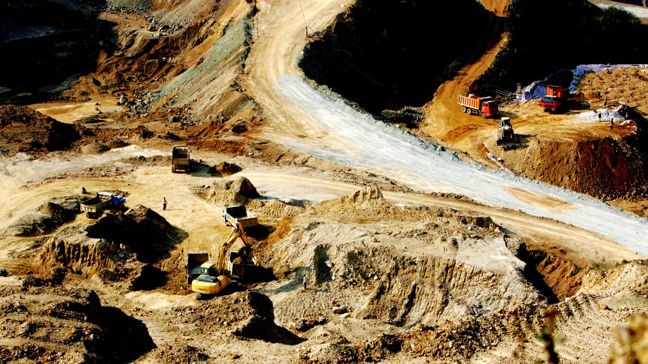 Un grande sito minerario a cielo aperto con escavatori, autocarri con cassone ribaltabile e operai che lavorano su un terreno irregolare e ricoperto di terra, sotto la luce diretta del sole.