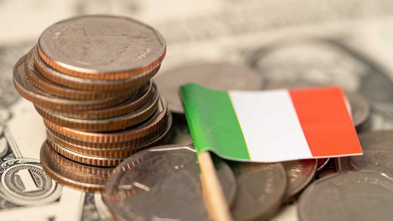 Una piccola bandiera italiana su uno stuzzicadenti è posta tra una pila di monete e un po' di carta moneta sullo sfondo. Le monete sono per lo più di rame e la carta moneta sembra essere dollari americani.