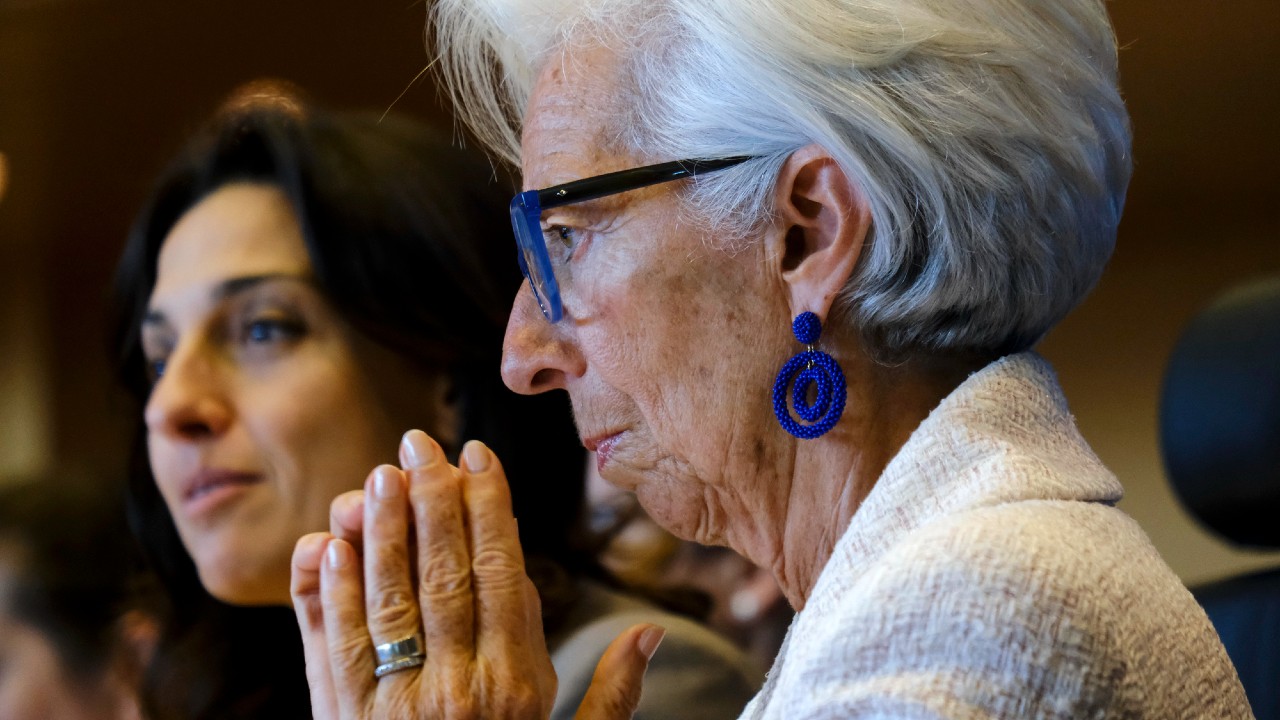 Due donne sedute vicine; quella in primo piano ha i capelli bianchi, indossa orecchini blu e occhiali, e tiene le mani giunte. La donna sullo sfondo ha i capelli scuri. Entrambe appaiono concentrate e impegnate in una conversazione o in un evento.