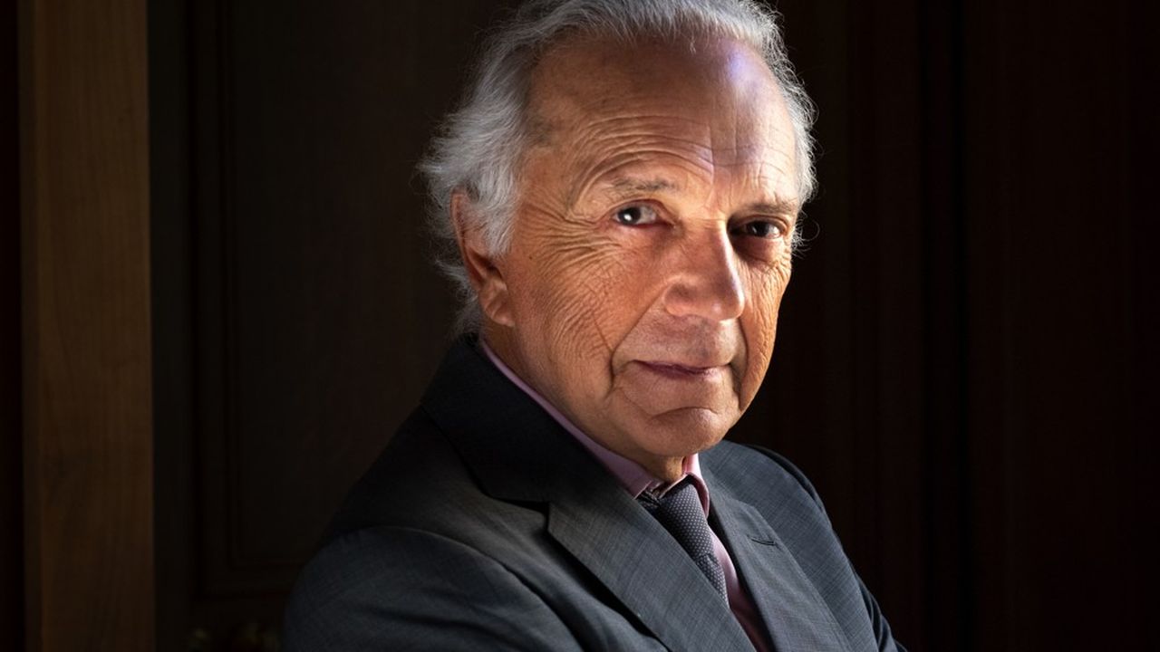 Un uomo anziano con i capelli grigi e in giacca e cravatta è in piedi con le braccia incrociate, con un sorriso sicuro e gentile. Lo sfondo è scuro, e il suo viso è messo in risalto da una luce soffusa.